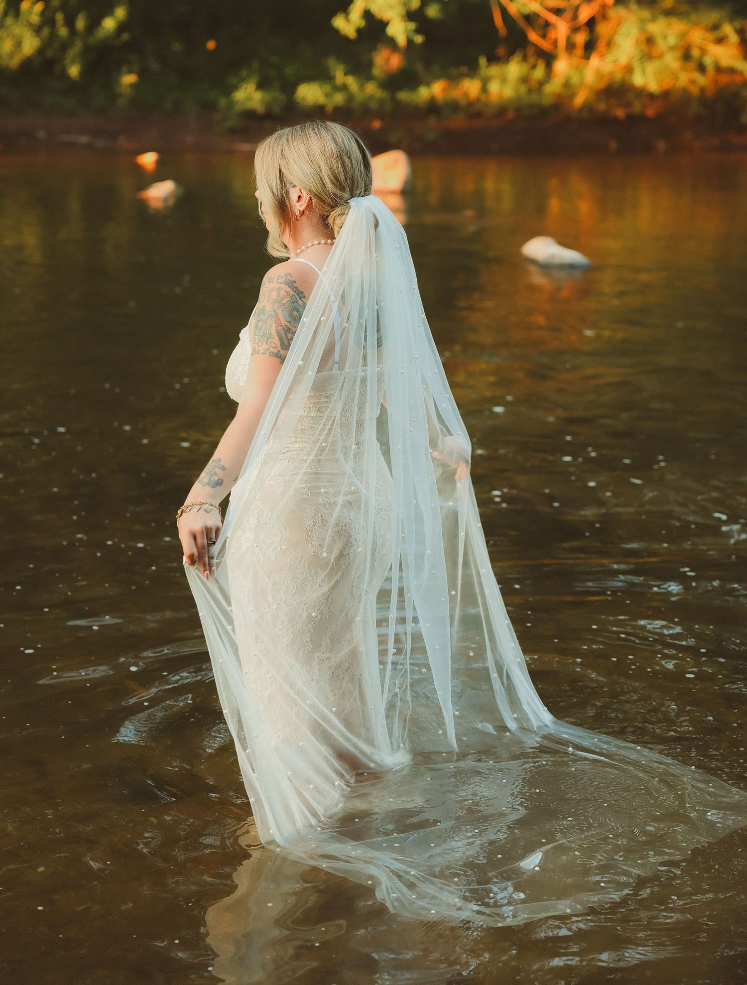 A flowing white wedding dress and veil create ethereal movement in glistening river water.