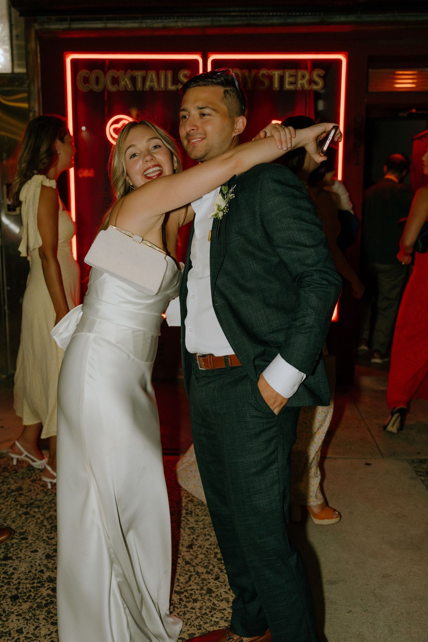 Wedding couple embracing and dancing beneath red neon signage at night.
