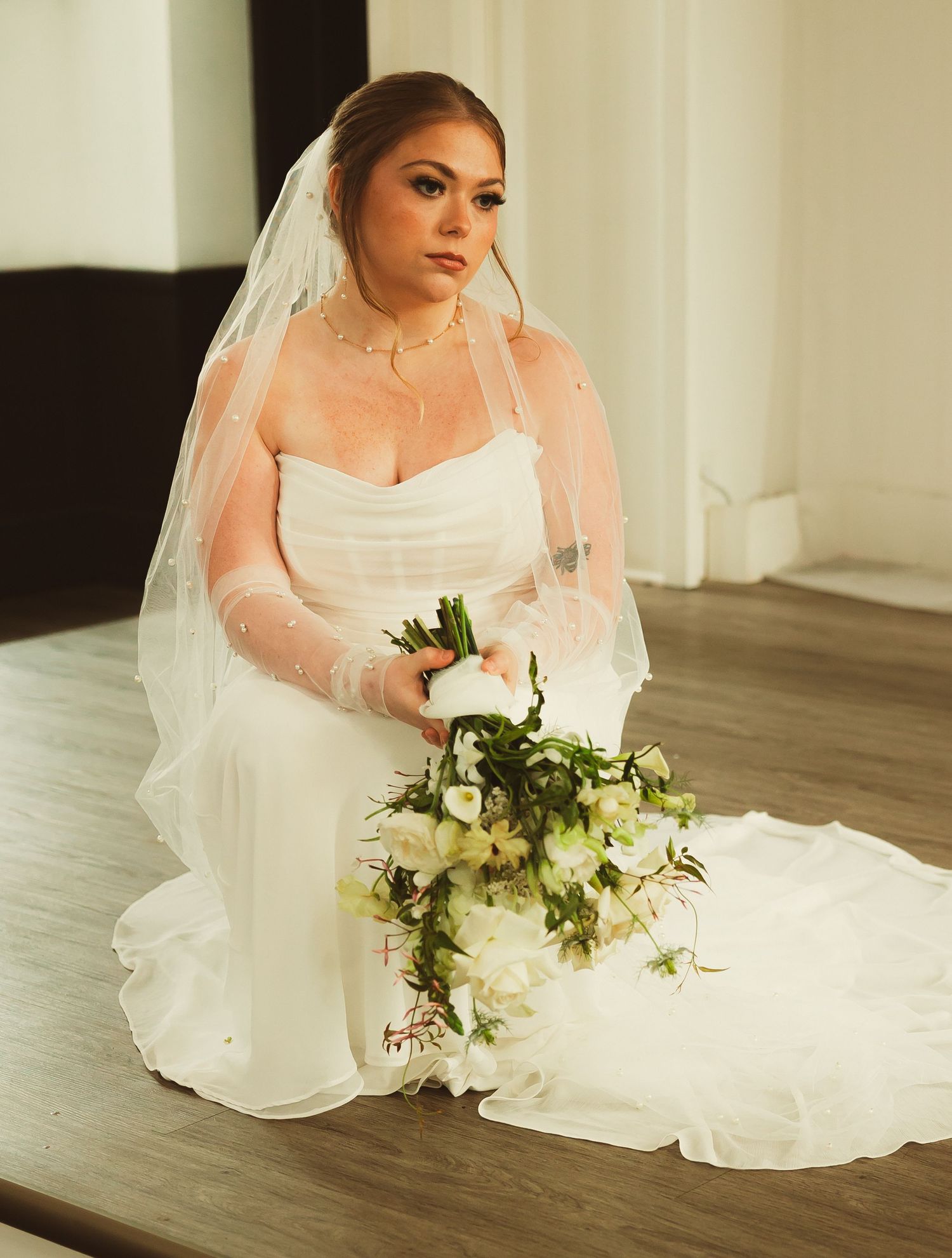 Bride seated on floor with white and green floral bouquet in natural light.