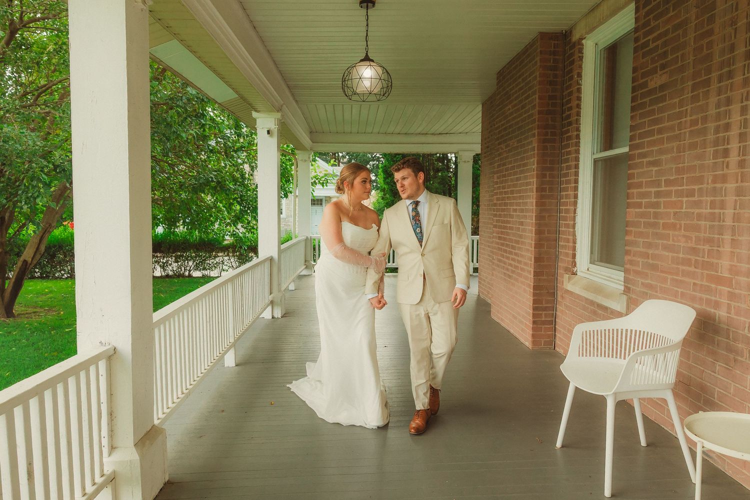 Newlyweds walking hand in hand along covered porch with white railing.