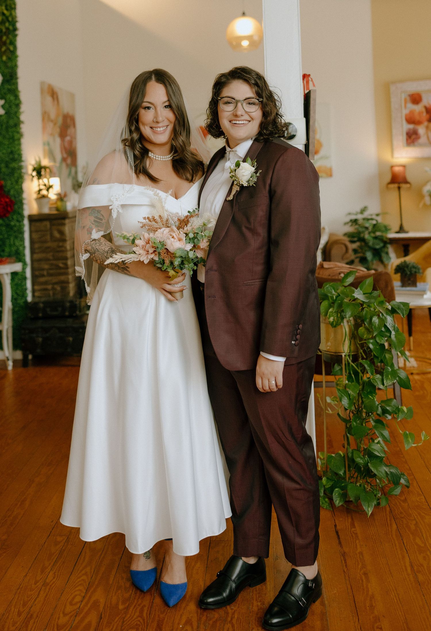 A couple poses together at their wedding, one wearing a white dress and the other in a brown suit with blue shoes.