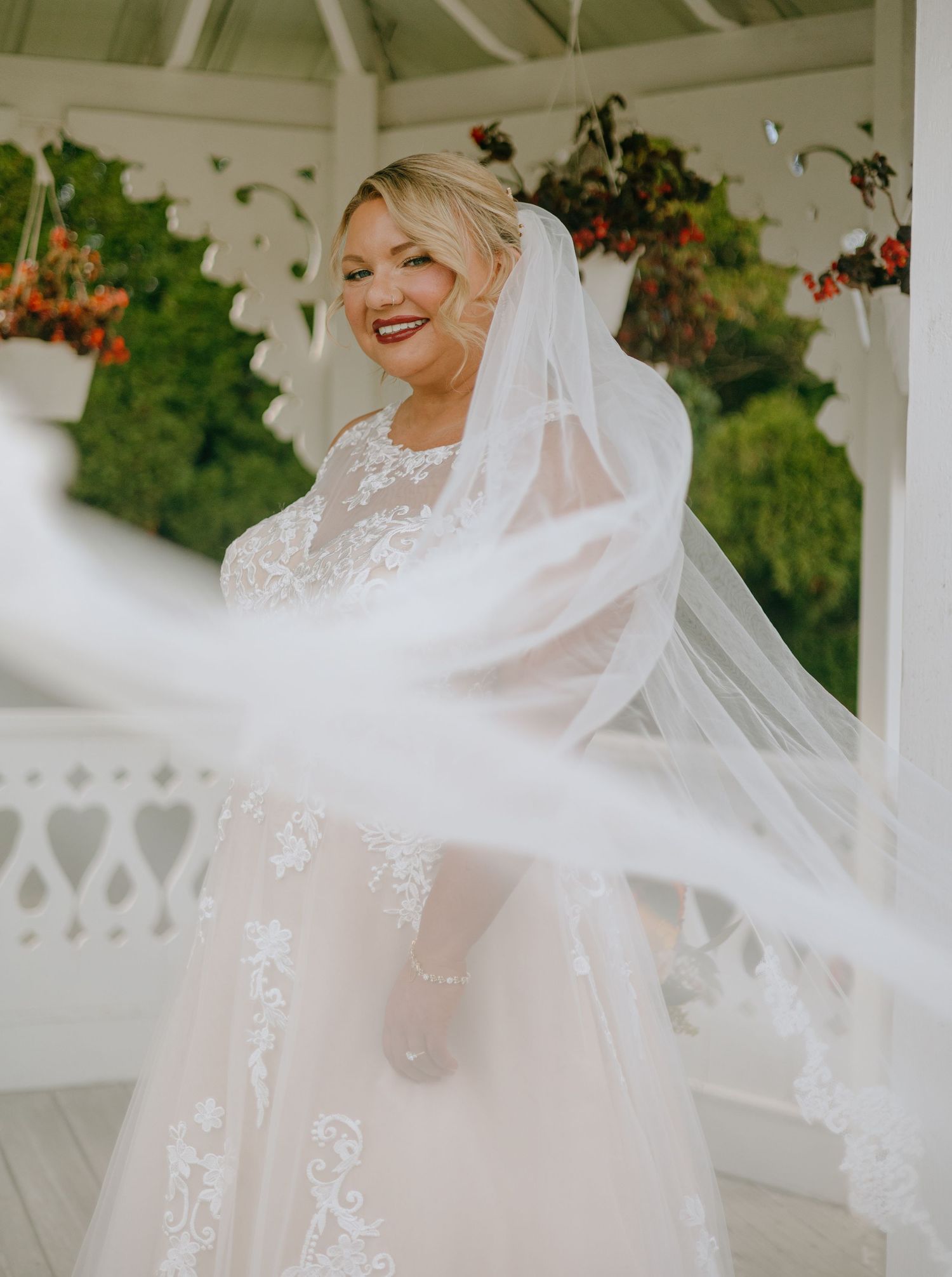 A bride in a flowing white lace wedding dress poses joyfully in a garden gazebo setting.