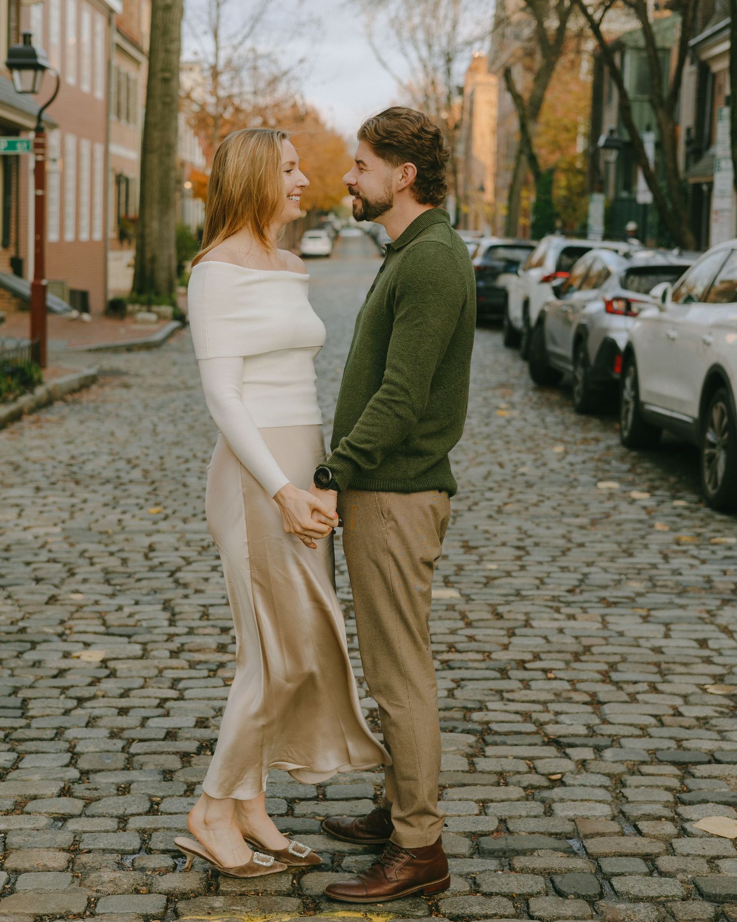 A couple shares a romantic moment on a historic cobblestone street lined with parked cars and autumn trees.