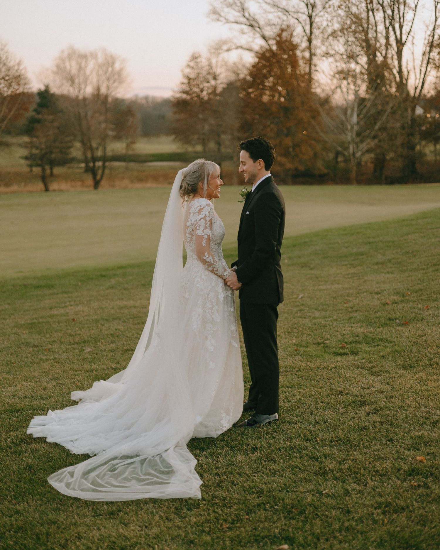 A wedding couple stands in a scenic field at sunset as the bride's long cathedral veil flows behind them.