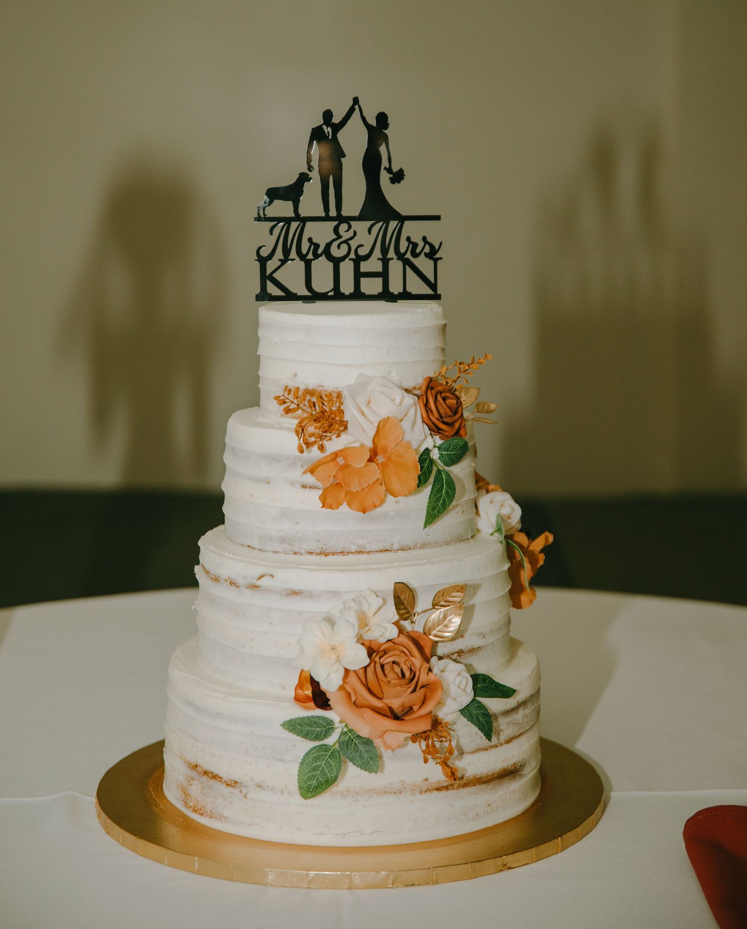Three-tiered white wedding cake decorated with peach roses and silhouette topper on gold base.