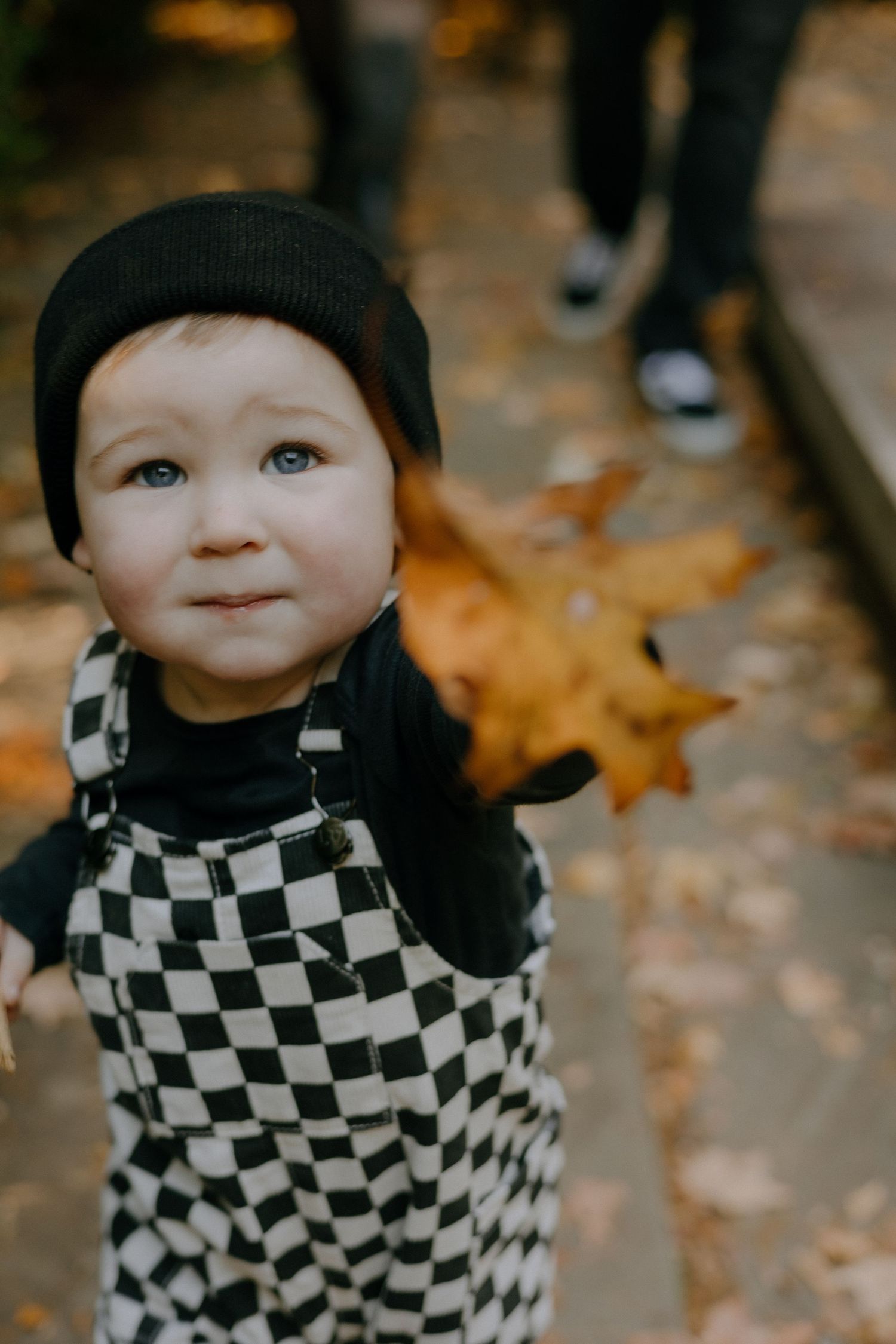 A small figure in a black beanie and checkered outfit smiles while playing outdoors in fall.
