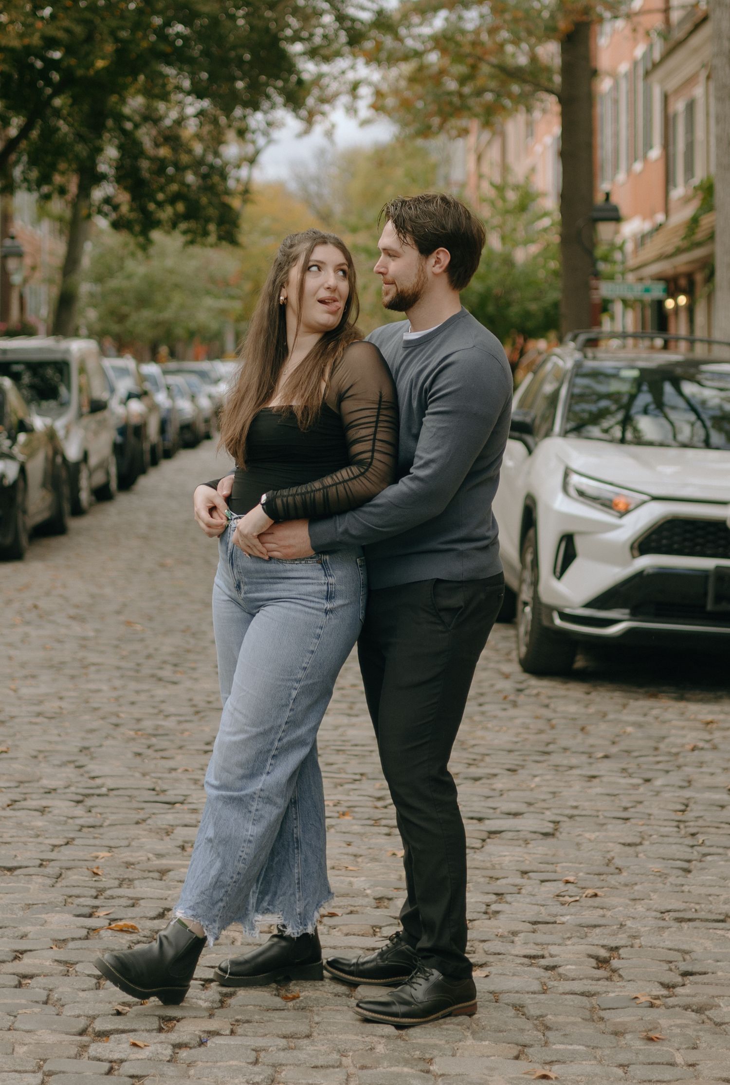 Romantic couple sharing playful moments on a tree-lined cobblestone street with parked cars.
