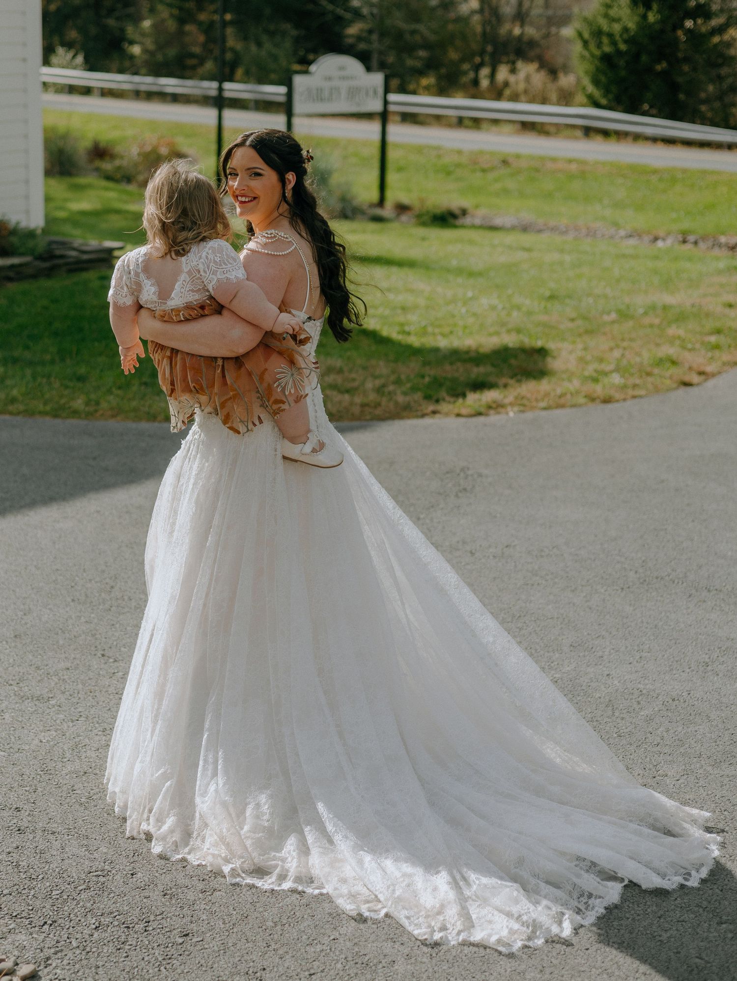 A bride in a flowing white gown holds a child on an outdoor path.