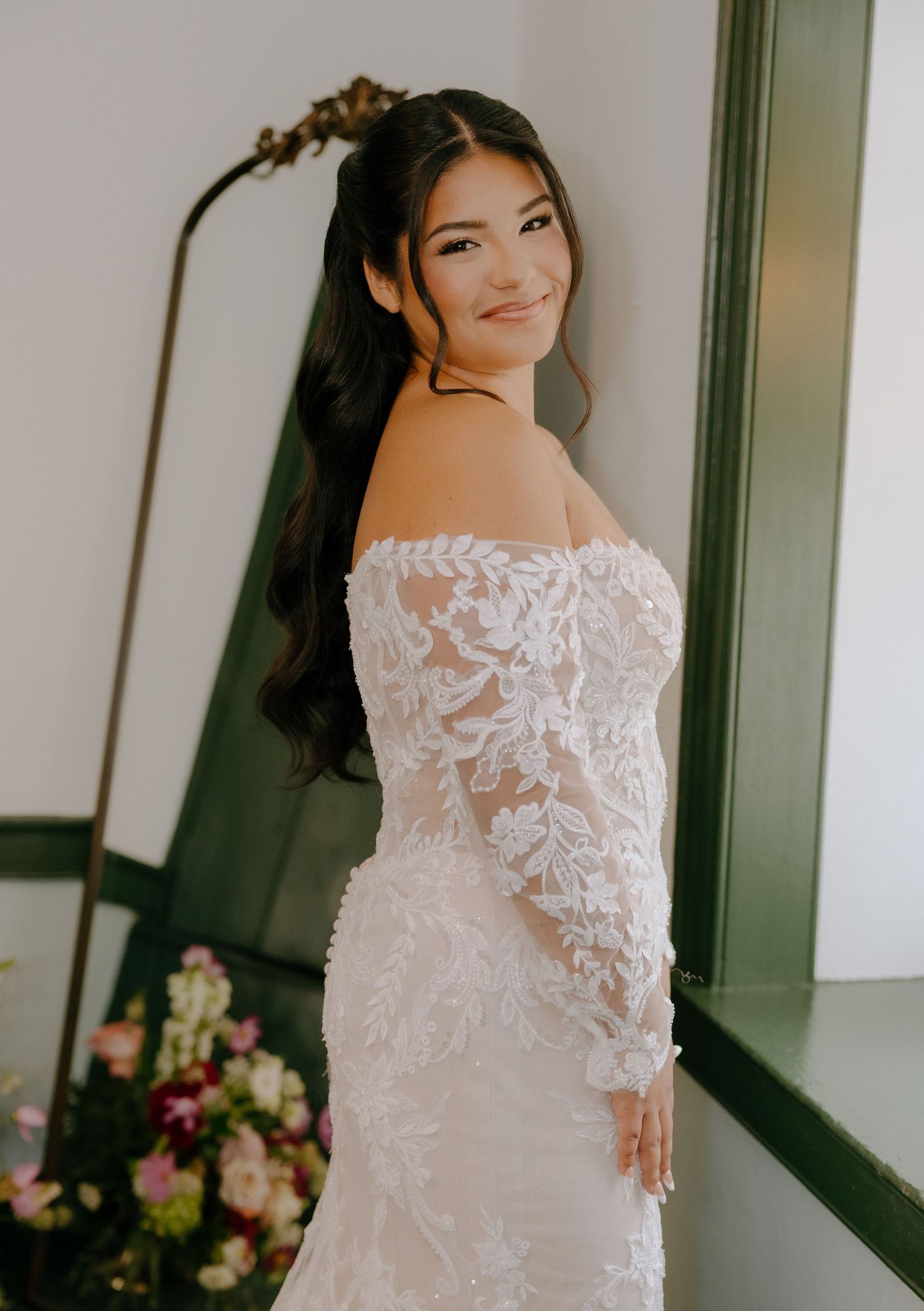 Bride in fitted lace gown smiles while posing against elegant interior.