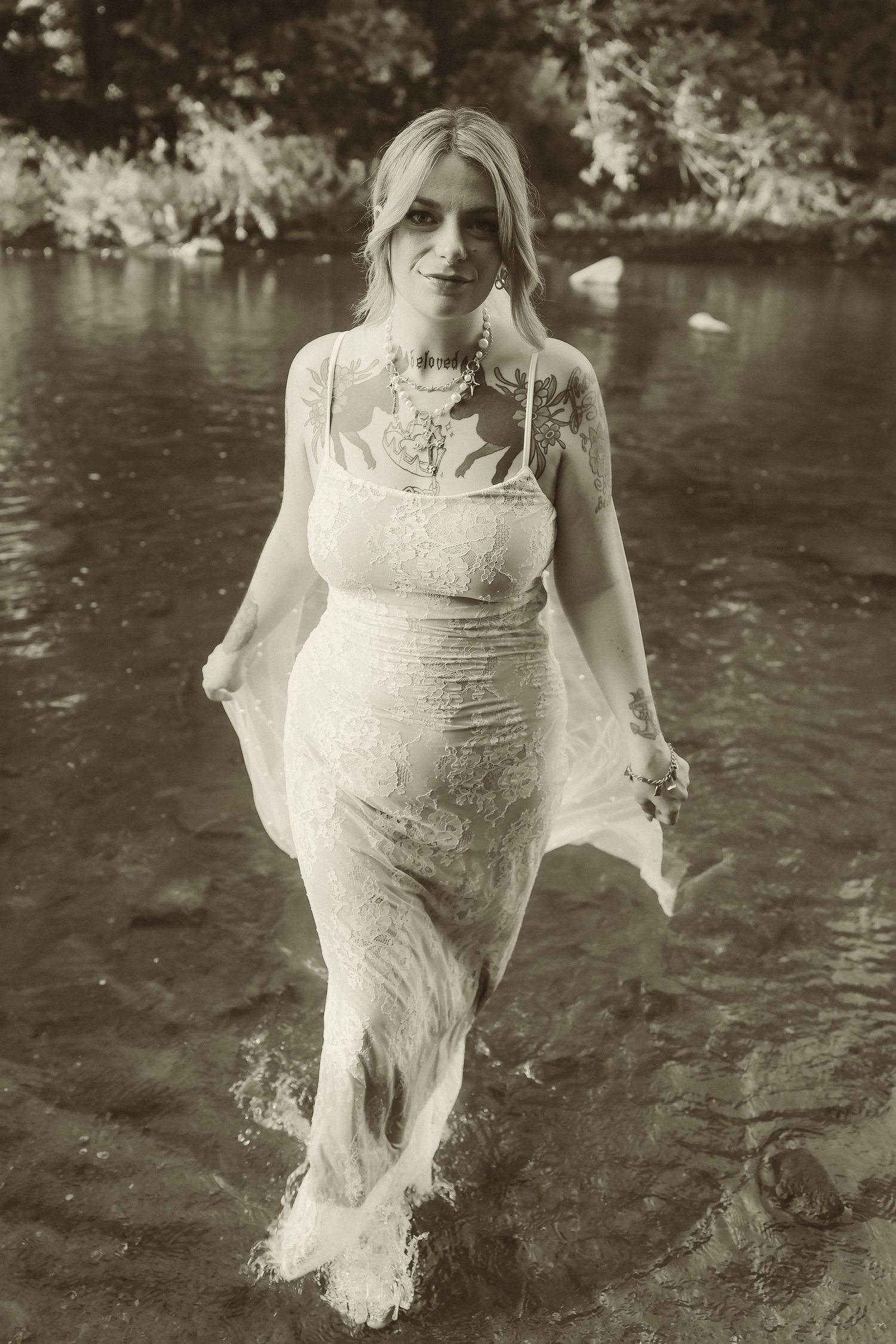 A black and white portrait of a bride walking through river water in her wedding dress.