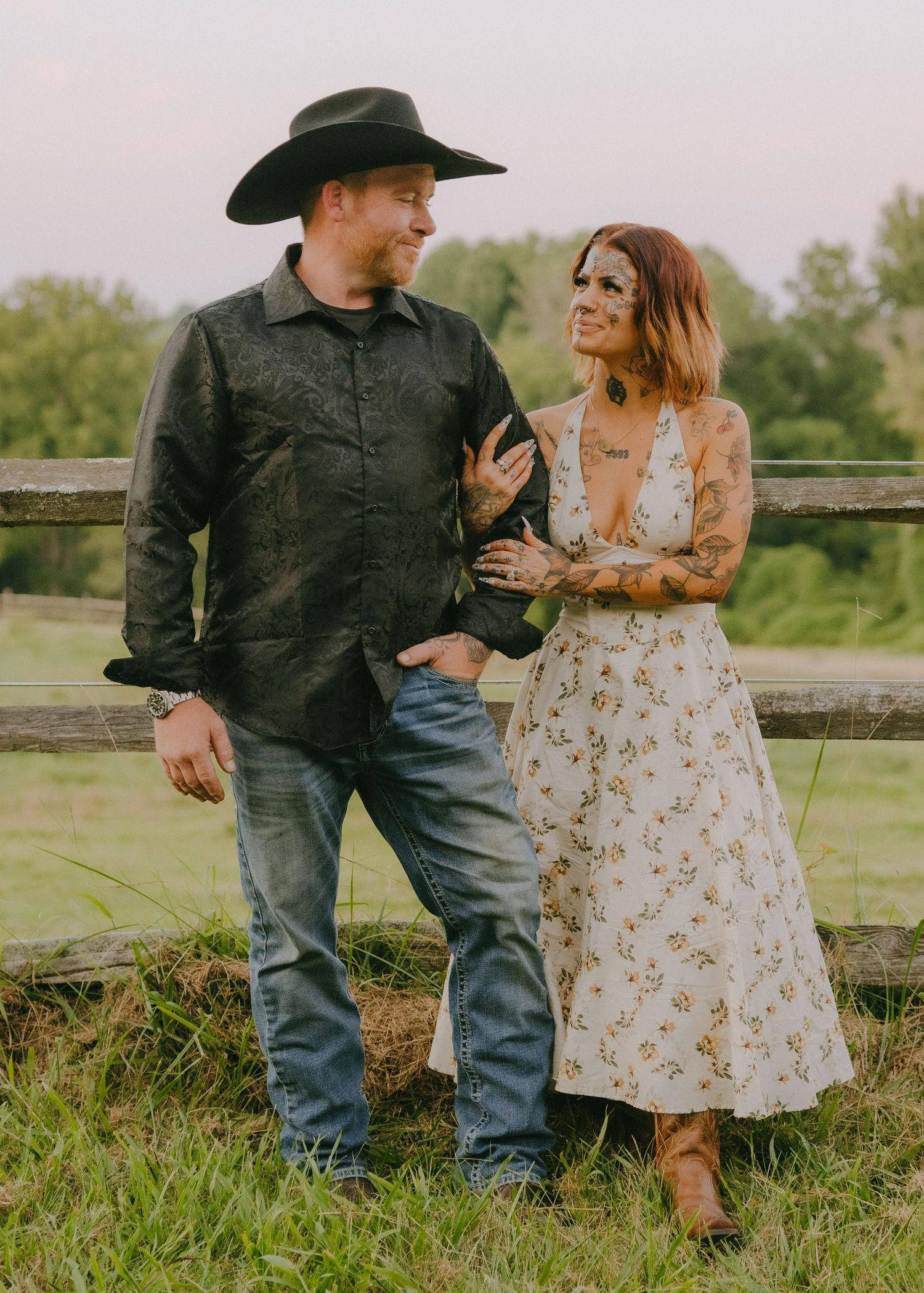 Couple shares intimate moment by rustic fence during countryside photo session.