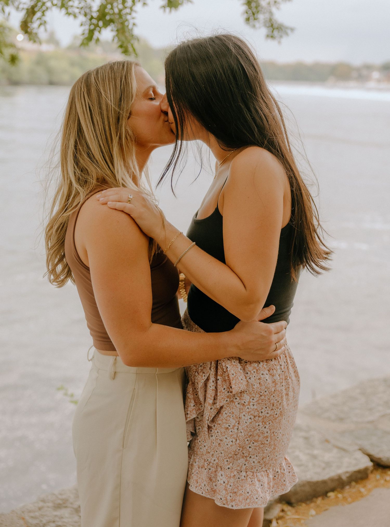 Two people sharing an intimate moment by a waterfront with trees in the background.