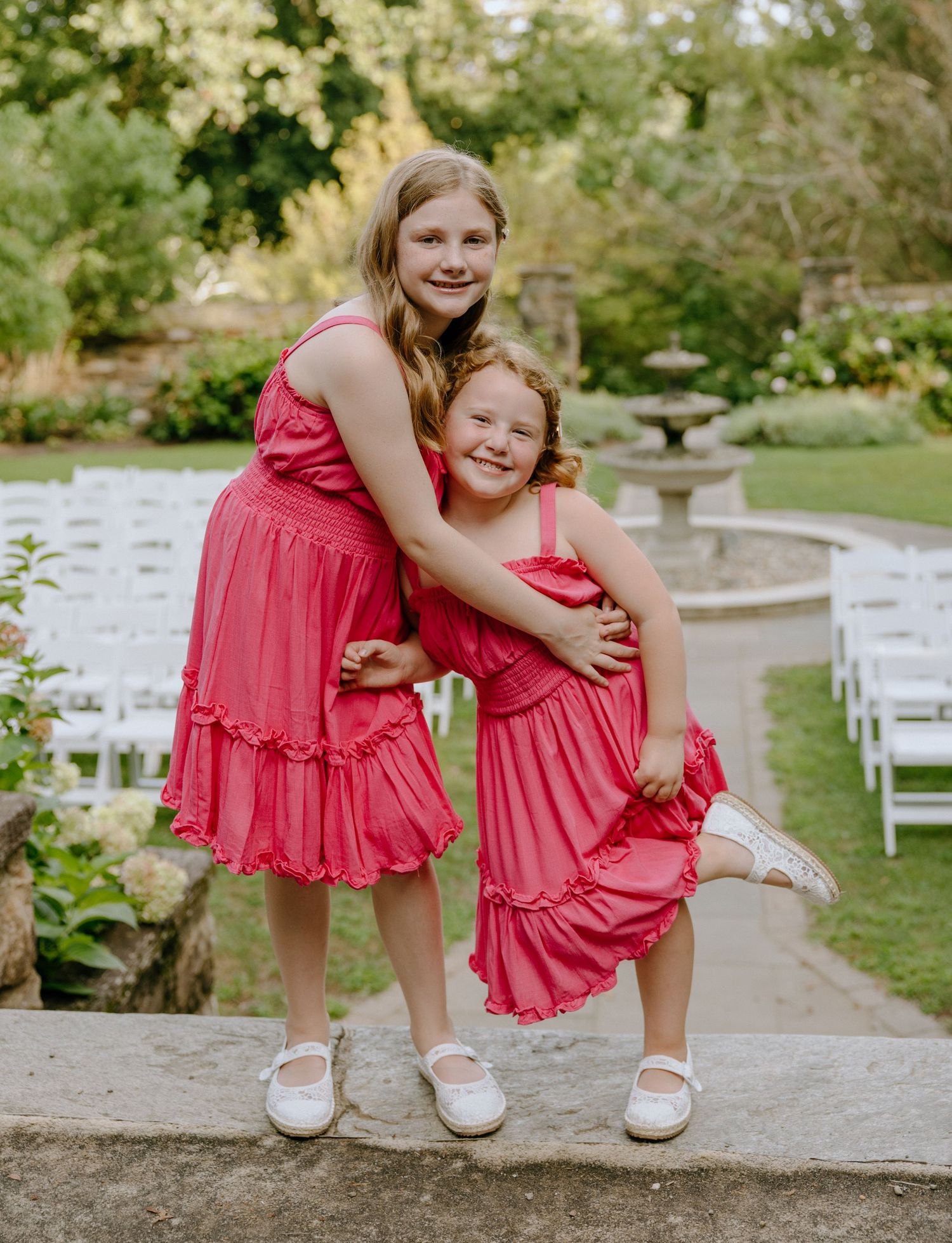 Two people wearing matching coral pink dresses pose playfully on garden steps with white chairs in background.