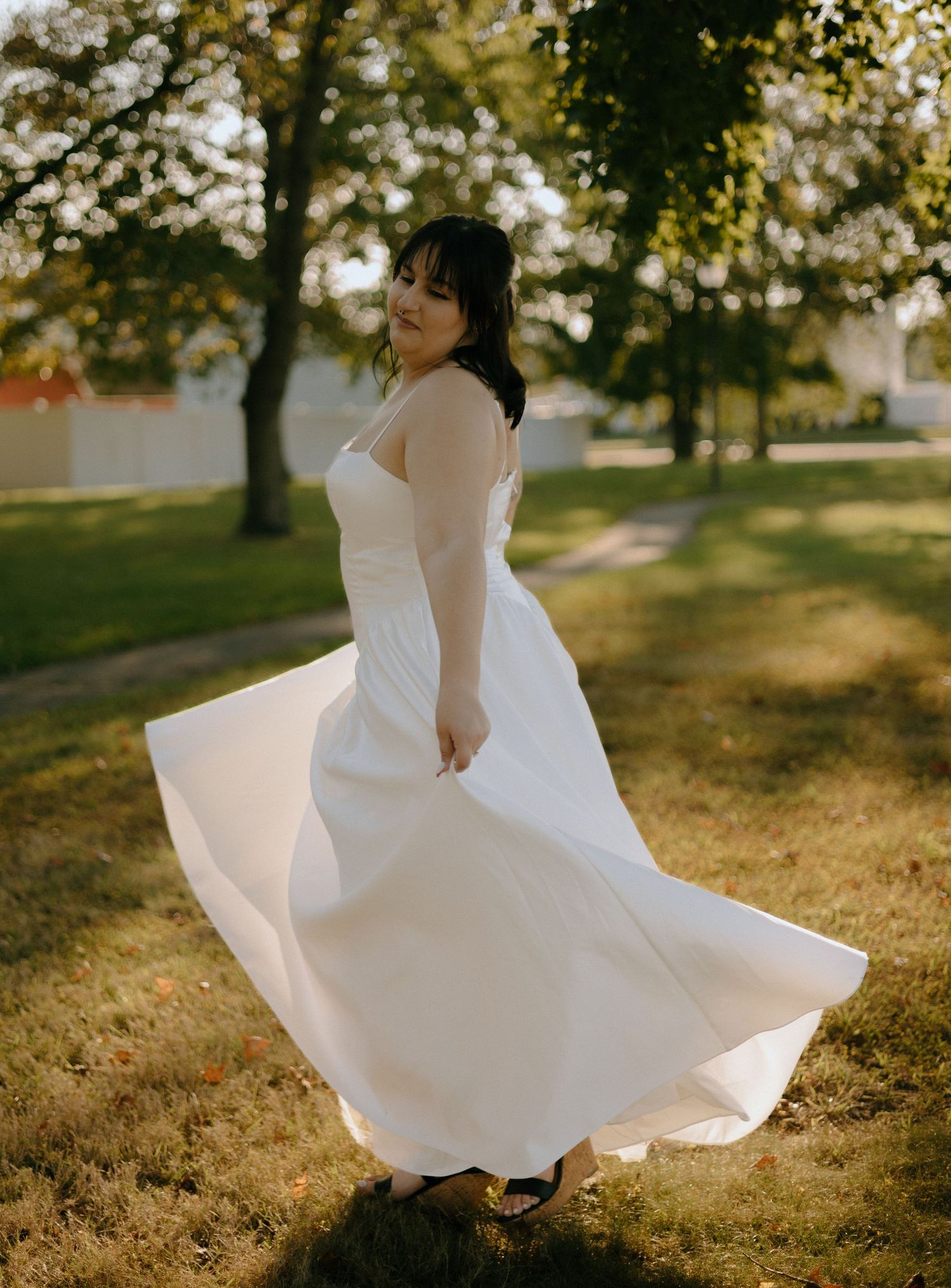 A bride in a flowing white dress twirls in a sunny park setting during autumn.