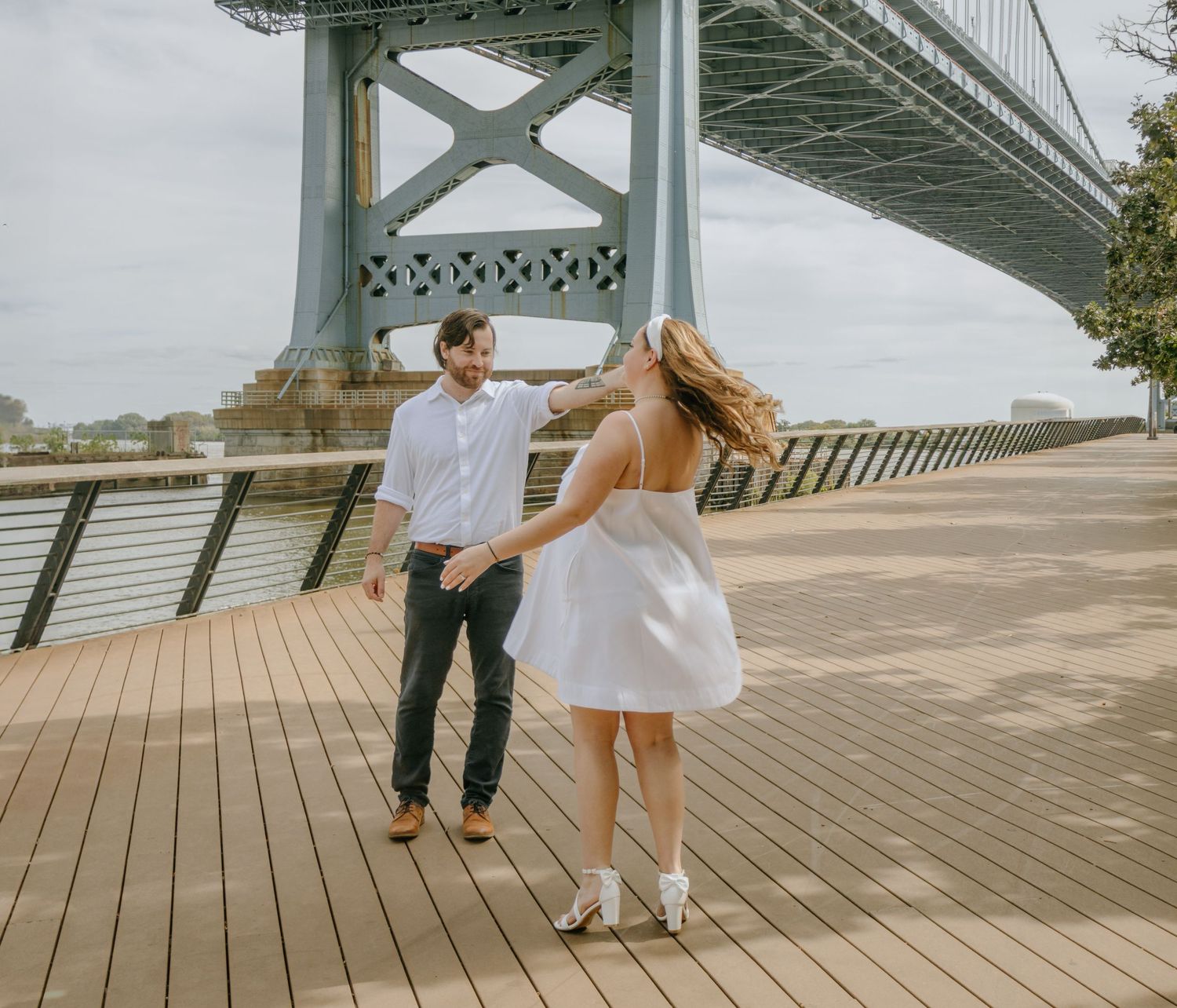 A couple stands on a wooden boardwalk under a bridge during a romantic engagement photoshoot.