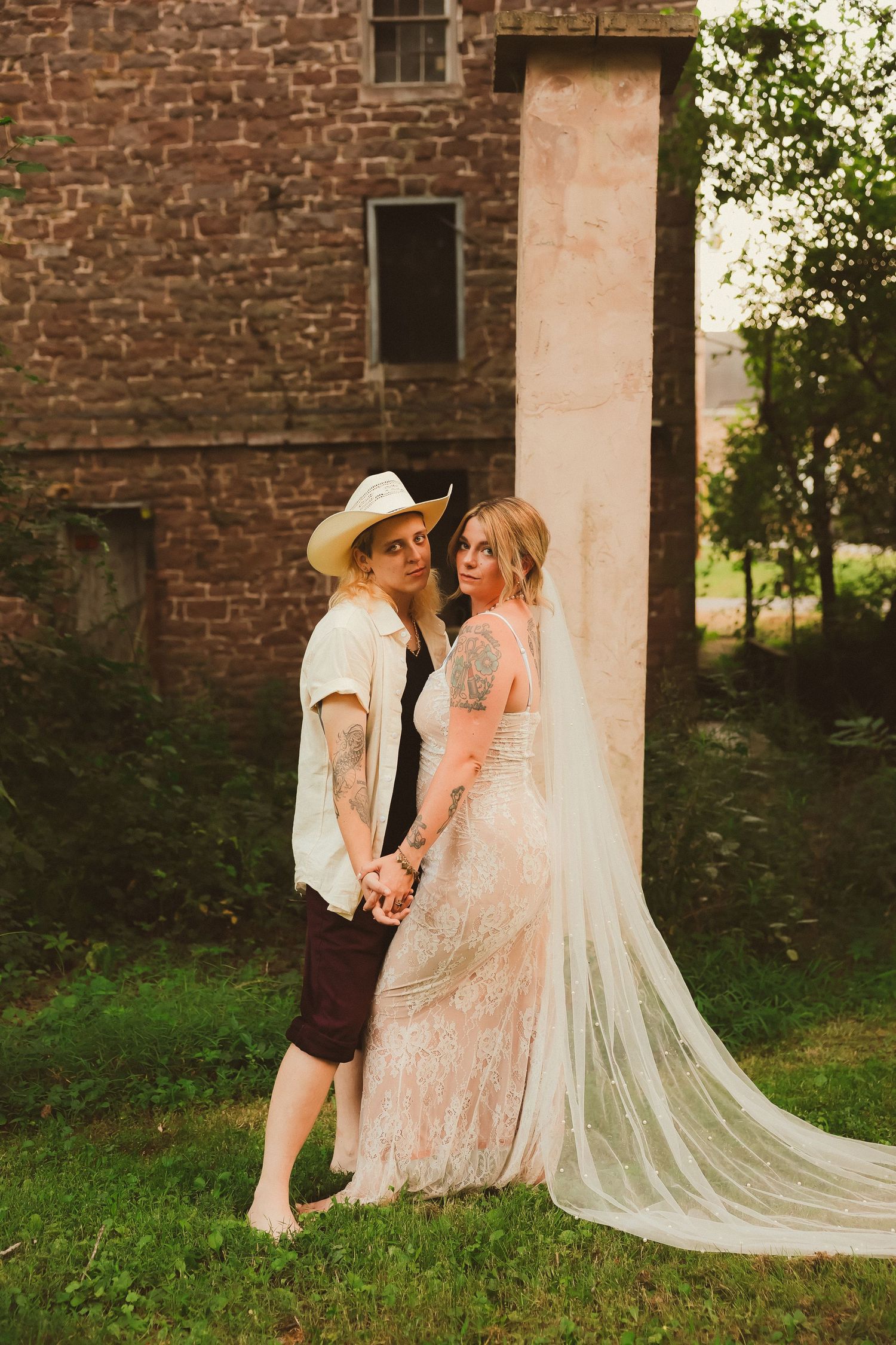 Two people in wedding attire share an intimate moment by a historic brick building at sunset.
