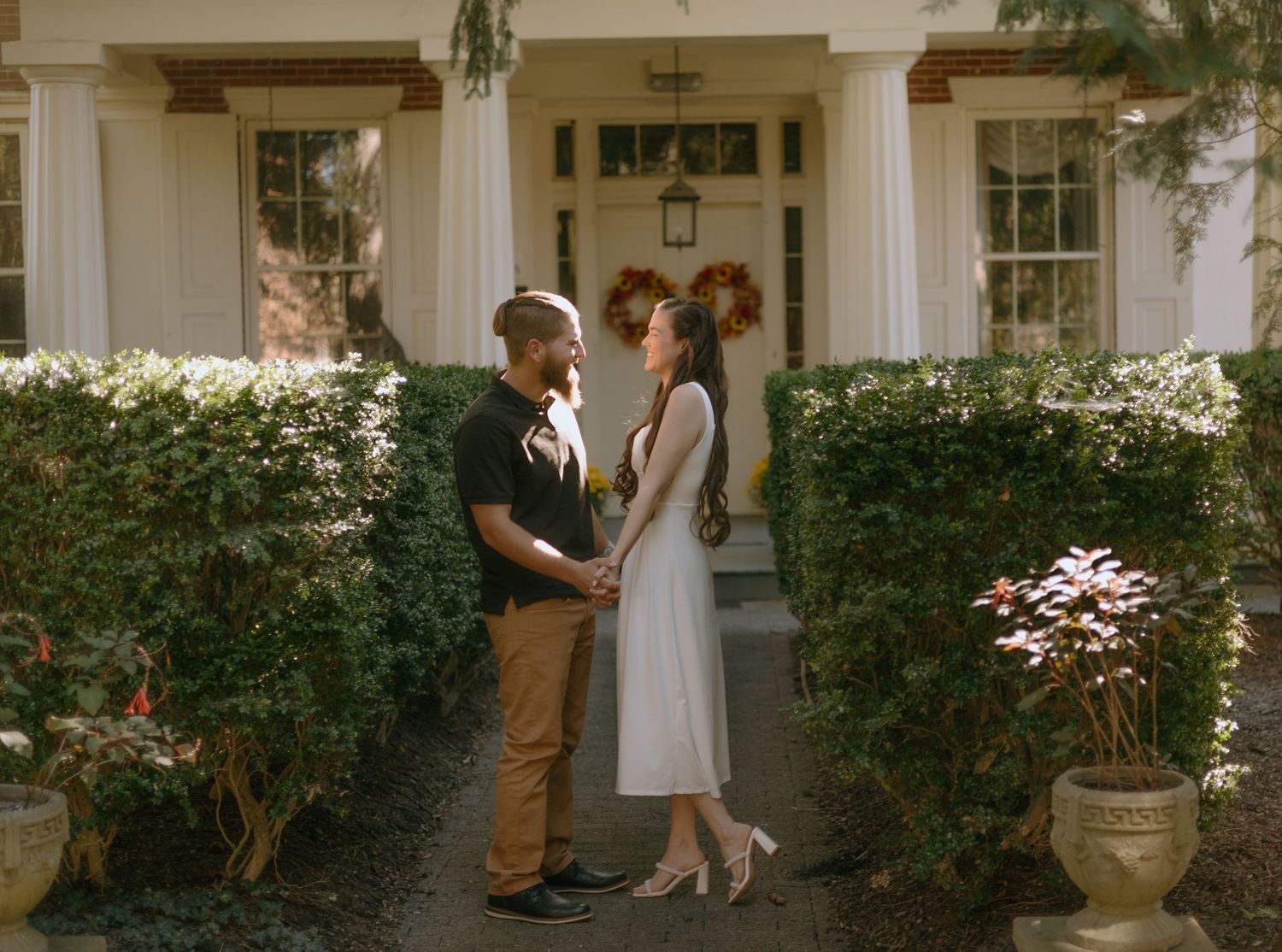 A romantic engagement photo series showing a couple in casual attire posing together in front of a white house exterior.