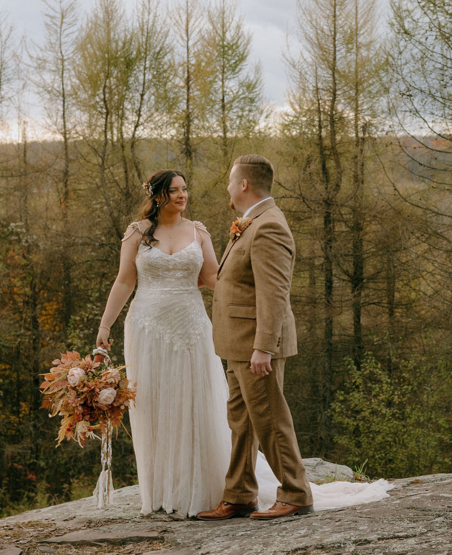 A couple in wedding attire sits together on a large rock formation with a scenic mountain backdrop.