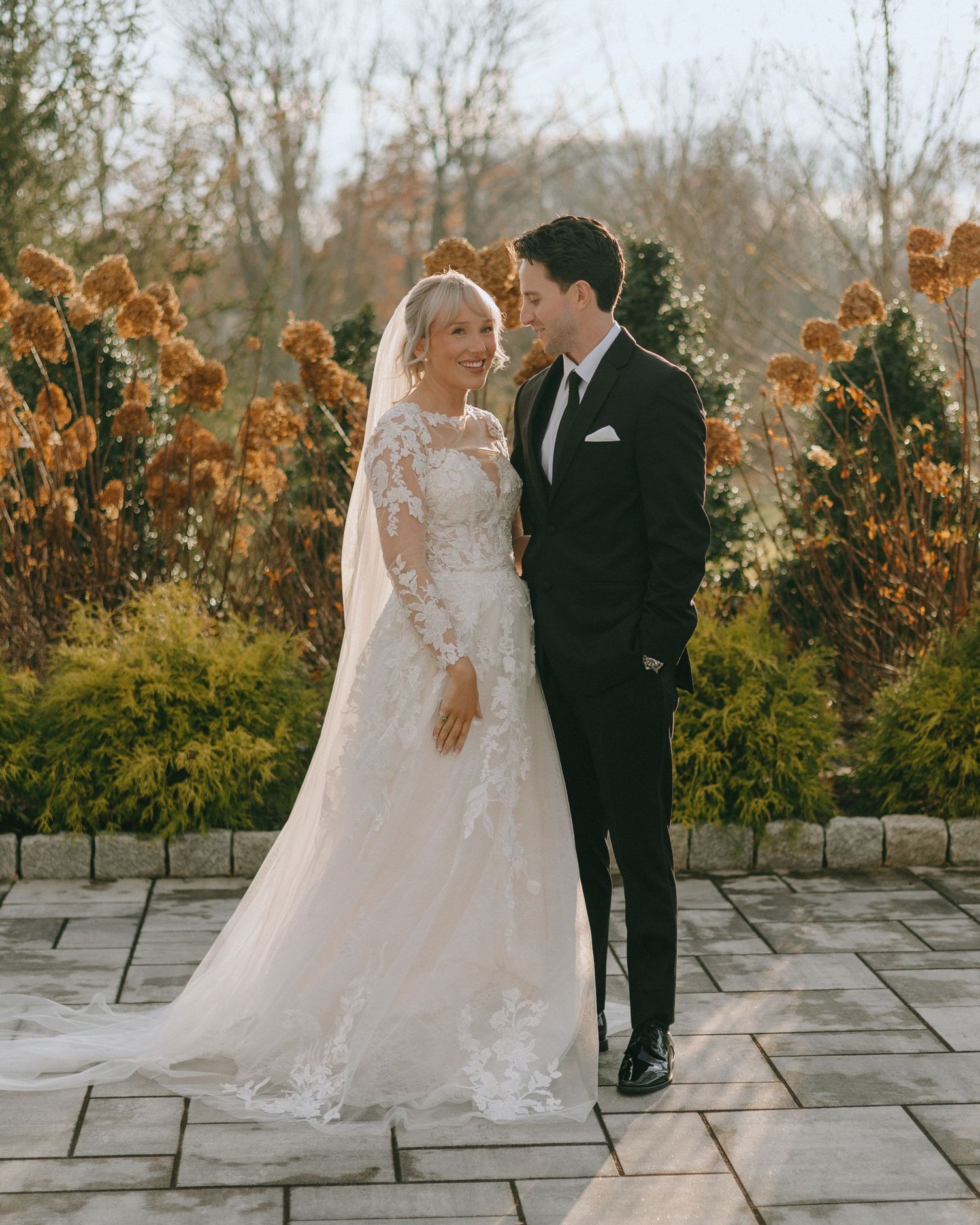 Romantic wedding couple poses for portraits on stone patio surrounded by autumn foliage at sunset.