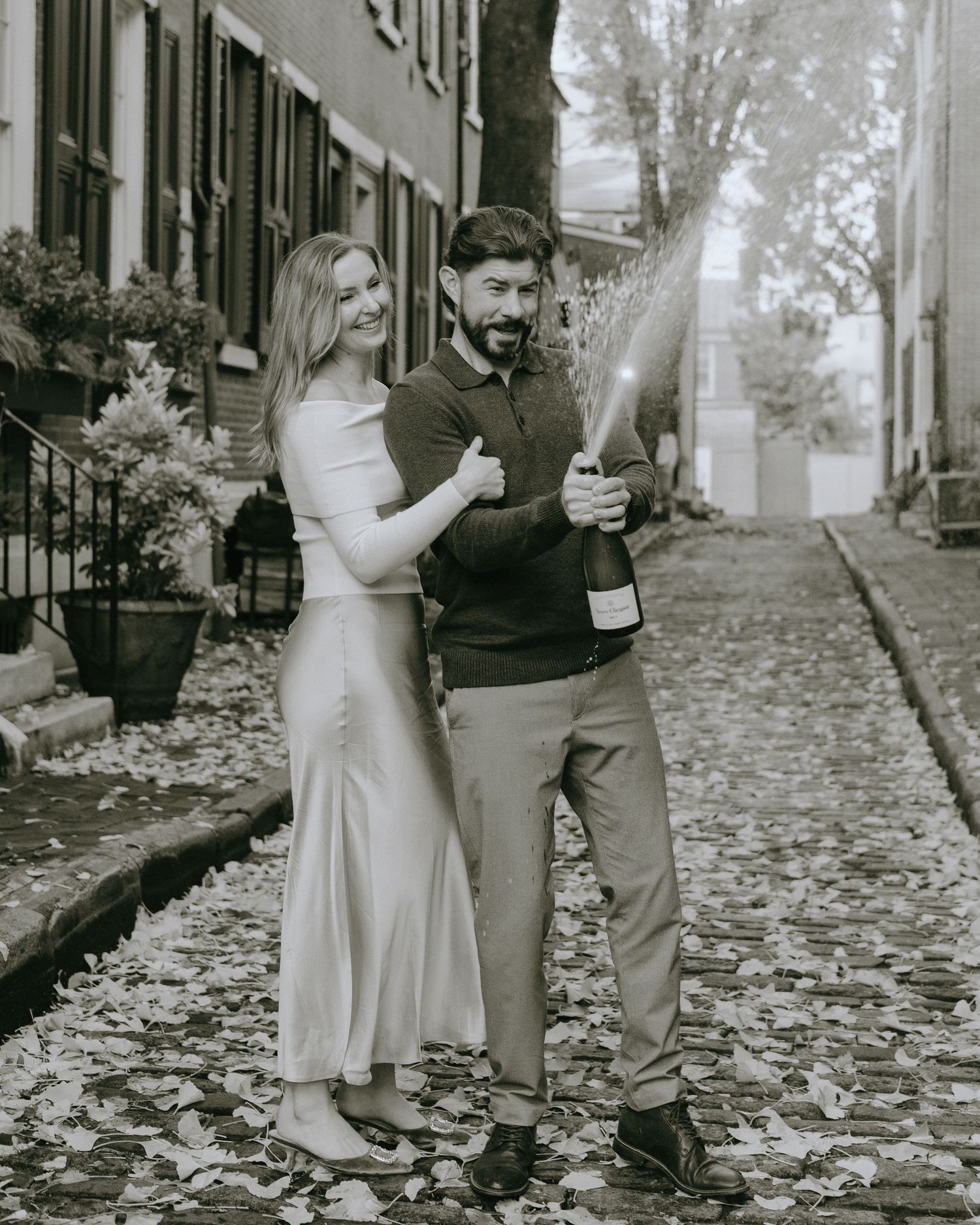Black and white photo of a couple popping champagne on a leaf-covered cobblestone street.