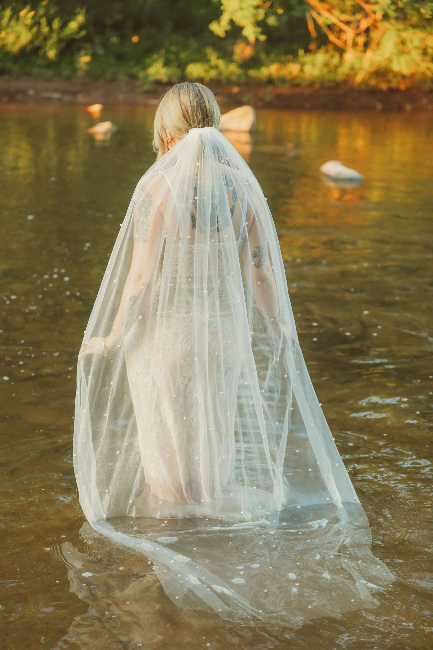 A figure in a flowing white wedding dress and veil stands in shallow water during golden hour.