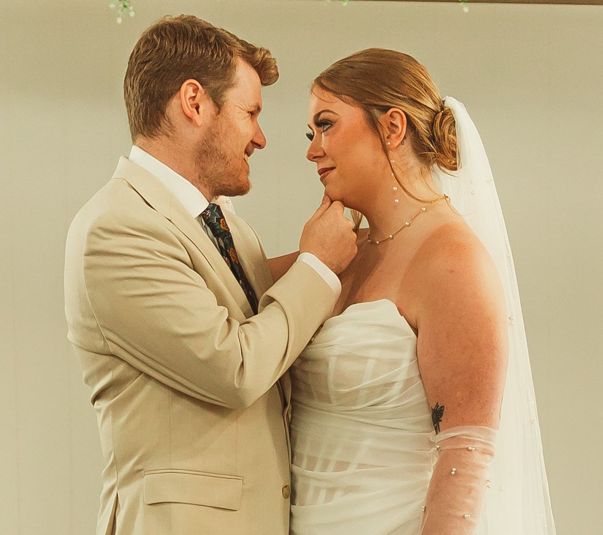 Close up of tender wedding day moment with couple in tan suit and white dress under greenery.