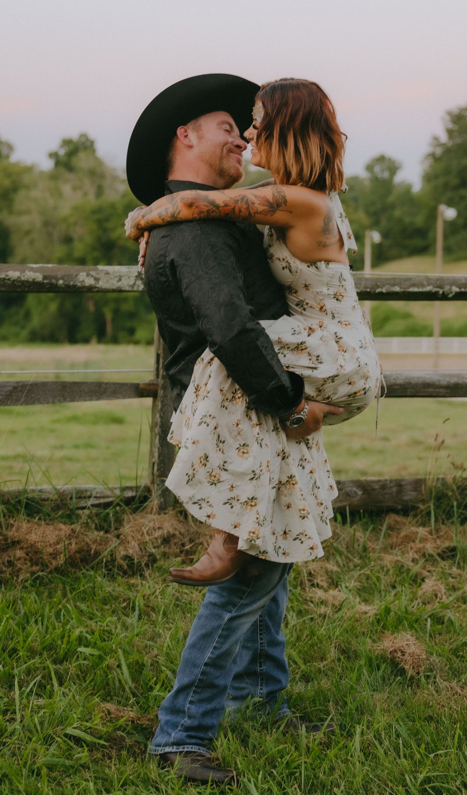 Two people embrace playfully by a wooden fence in a grassy field during golden hour.