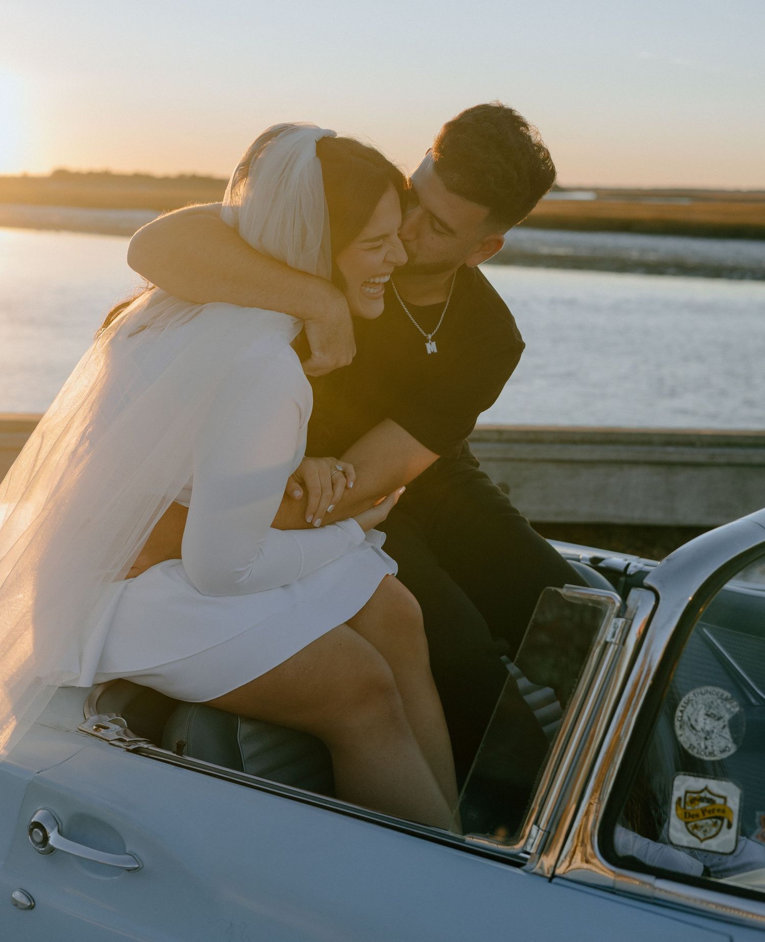 A couple shares an intimate moment in a vintage car during golden hour by the waterfront.
