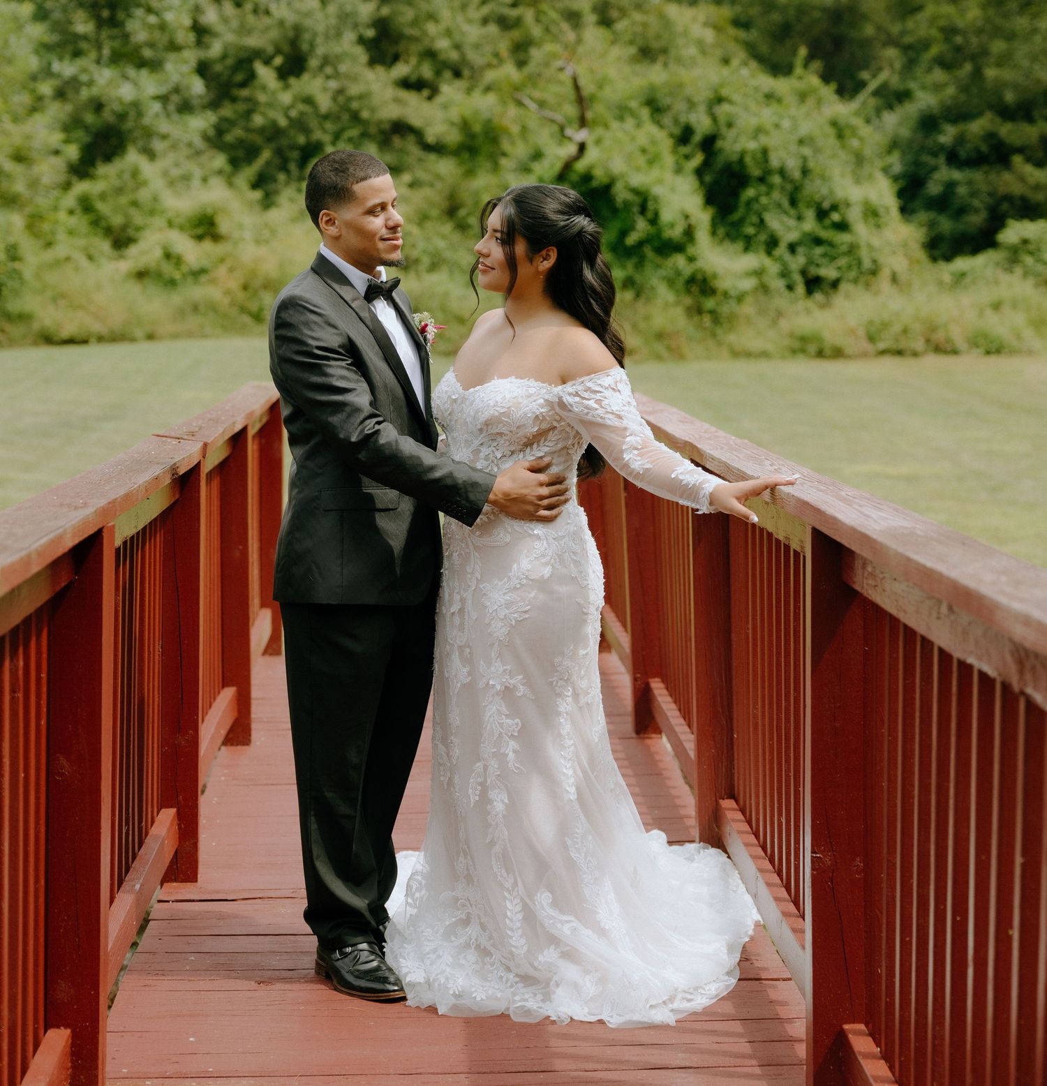Newlyweds share a romantic moment on a red bridge surrounded by greenery.