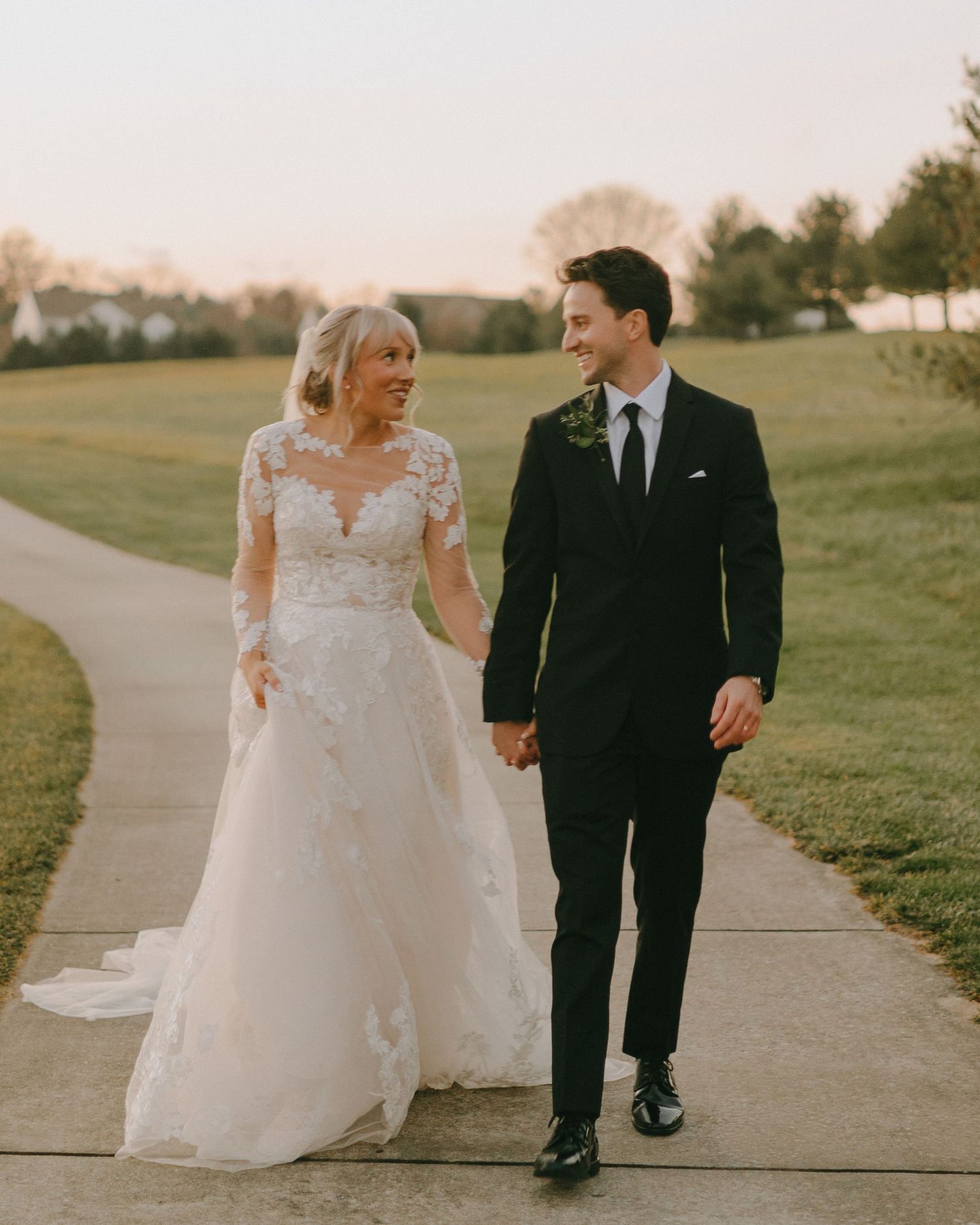 Newlyweds walk hand in hand along a path during sunset with rolling hills in the background.