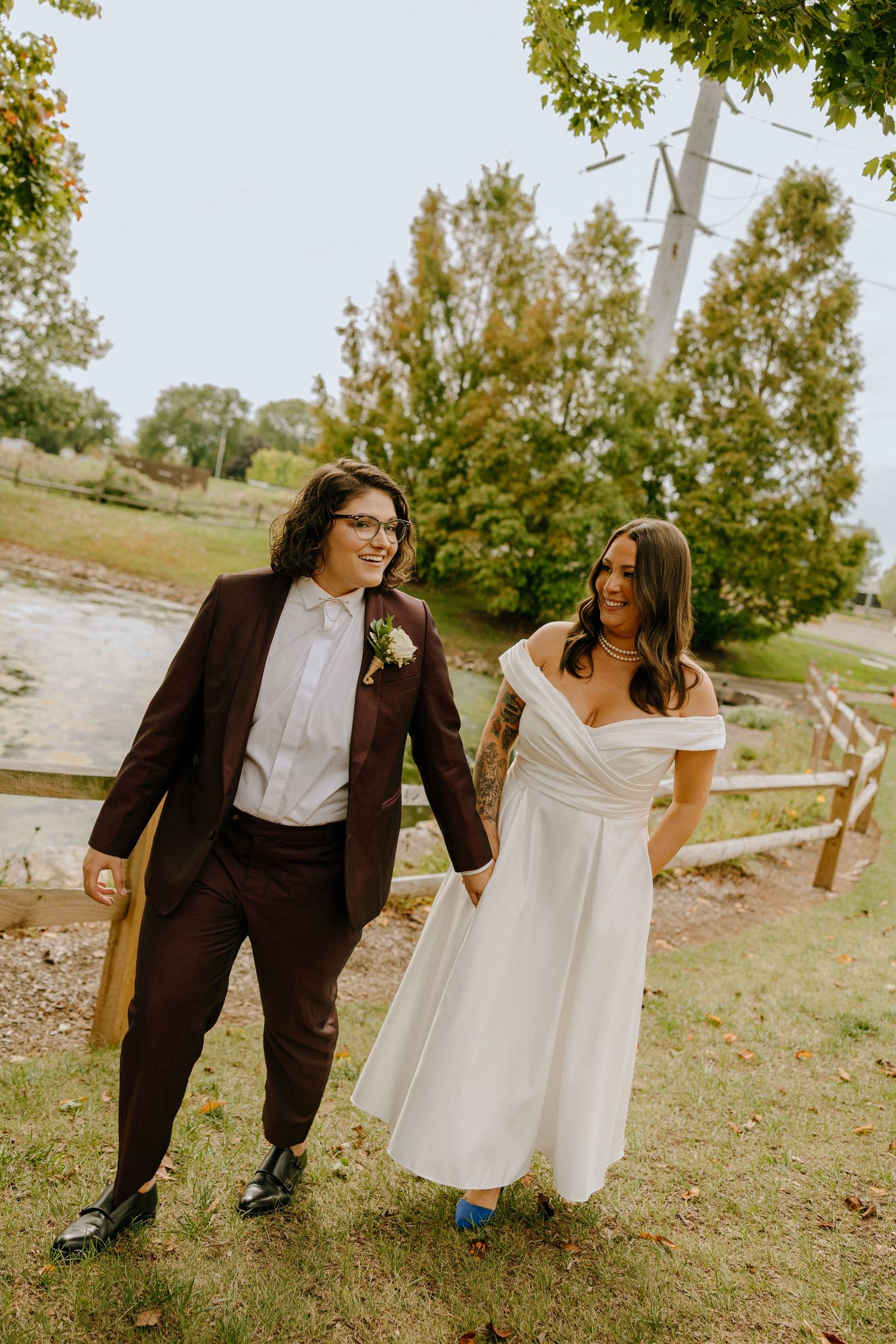 A couple walks together outdoors, one in a brown suit and one in a white dress with blue shoes.
