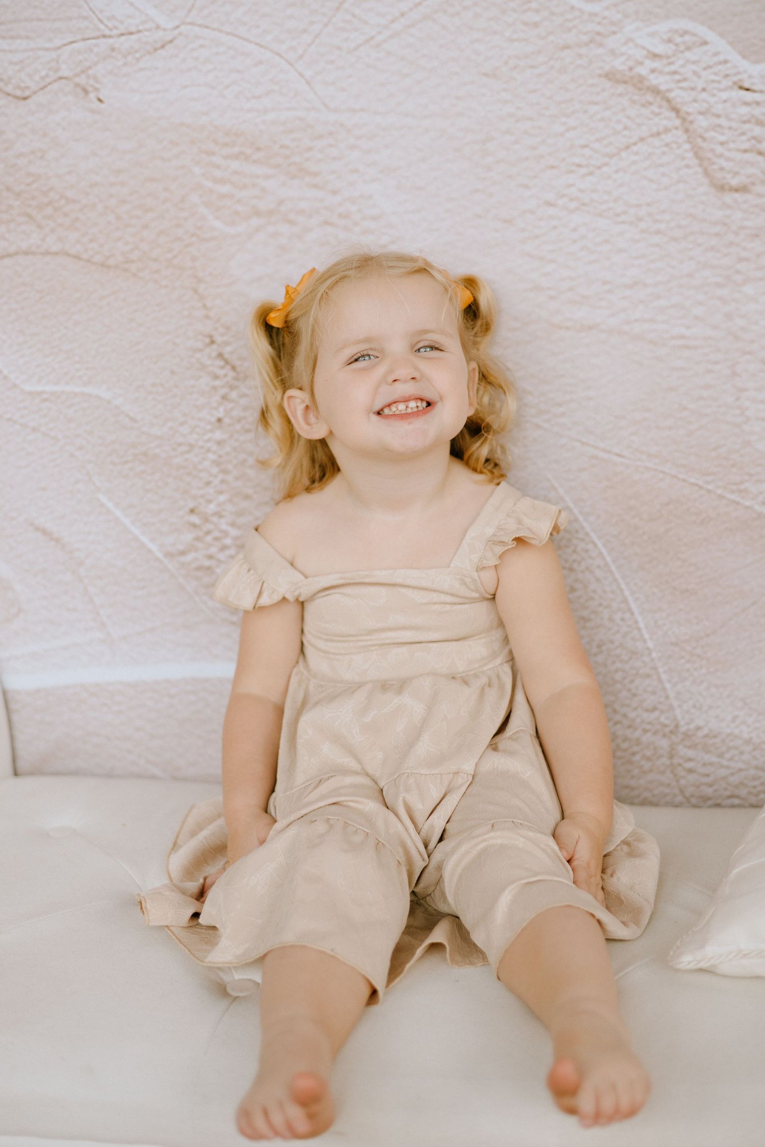 A small child wearing a beige romper sits smiling on a white couch with bare feet.