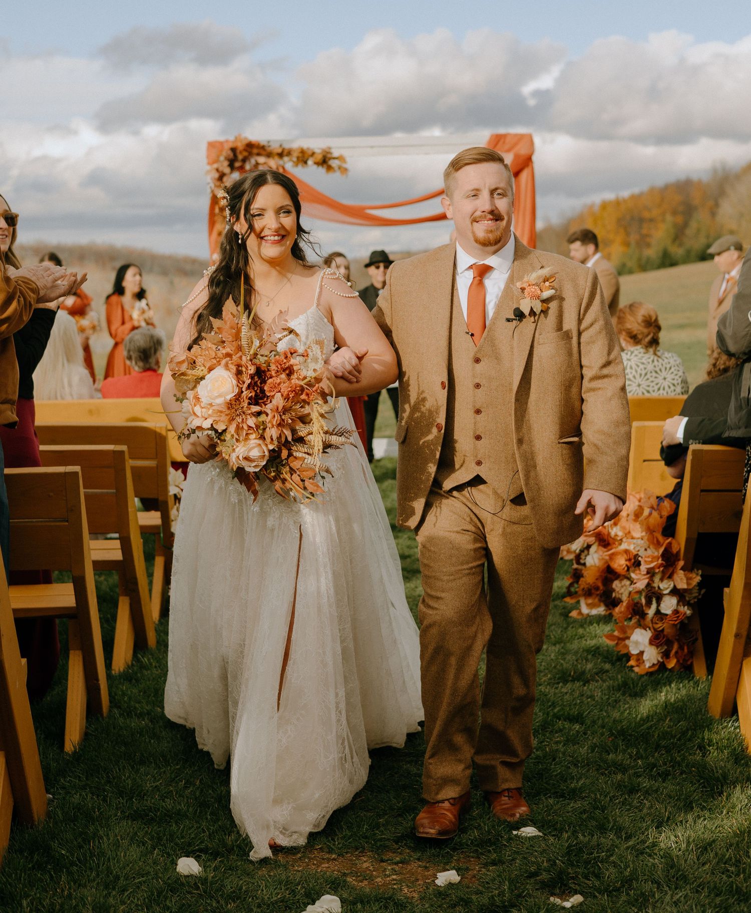 A wedding couple walks down the aisle at an outdoor autumn ceremony with wooden chairs.
