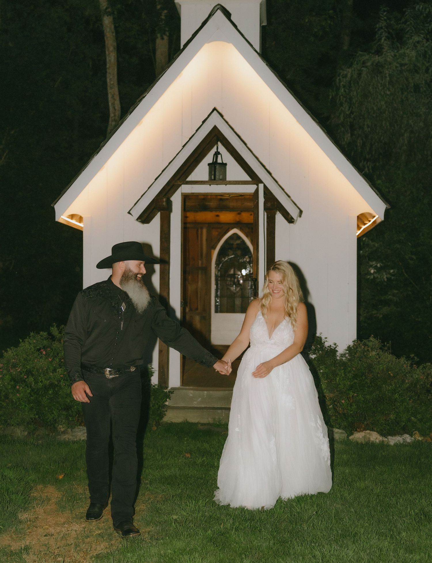 Night photos of newlyweds outside an illuminated white chapel in a garden setting.