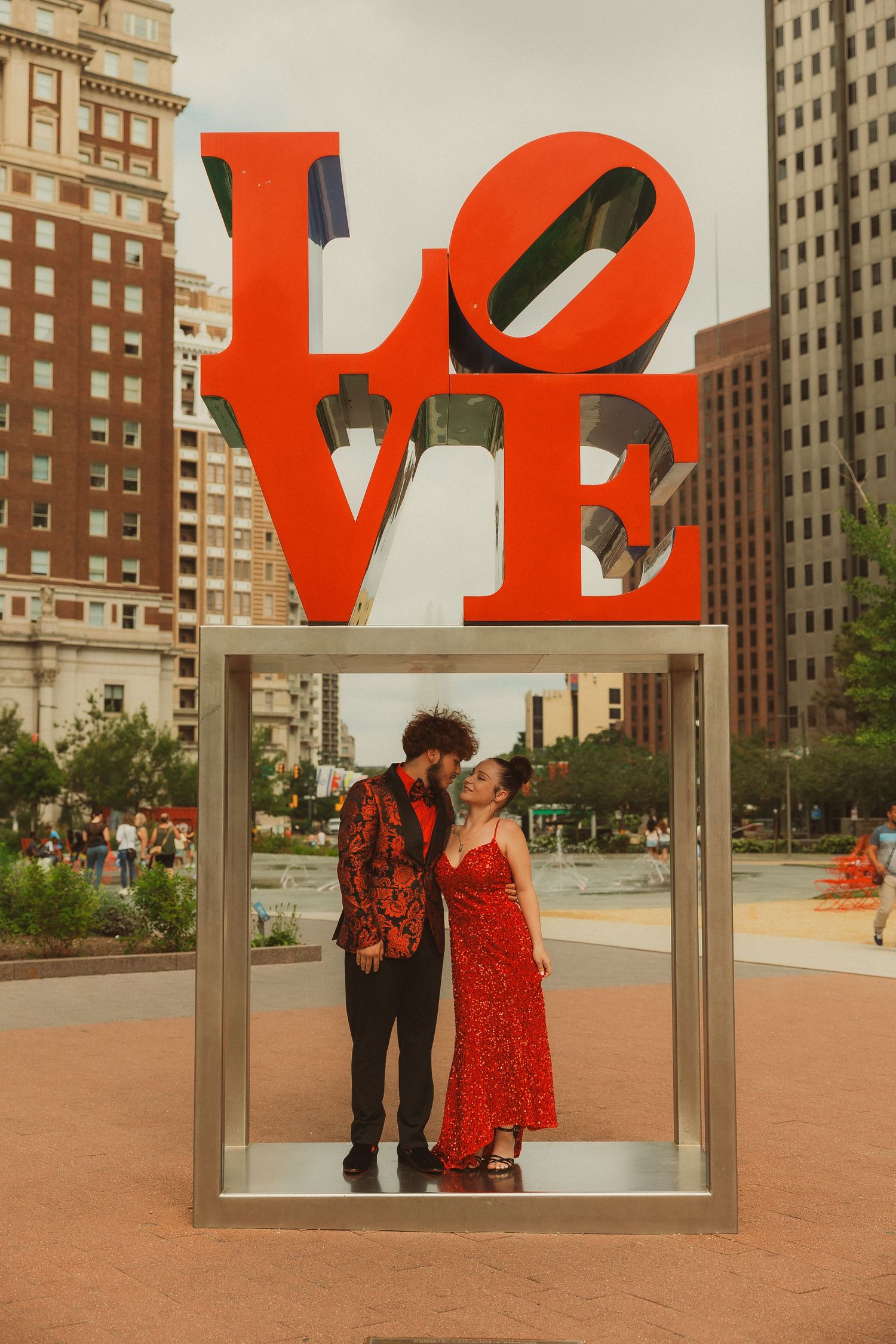 A couple poses inside a red LOVE sculpture installation in an urban plaza.