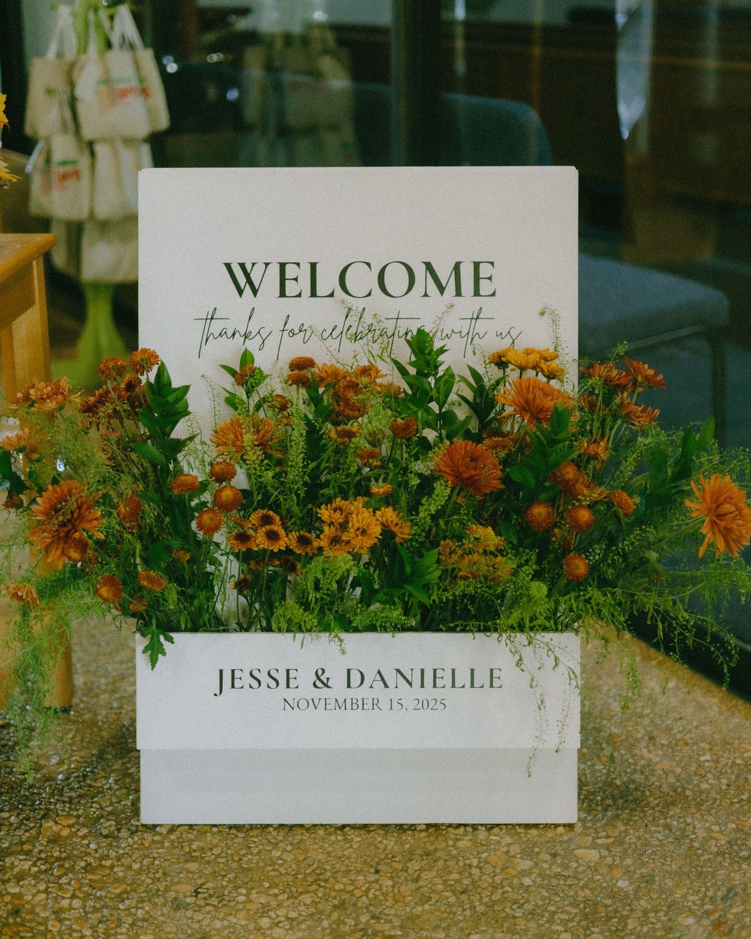 Wedding welcome sign with orange flowers and greenery displayed outdoors on a gravel surface.