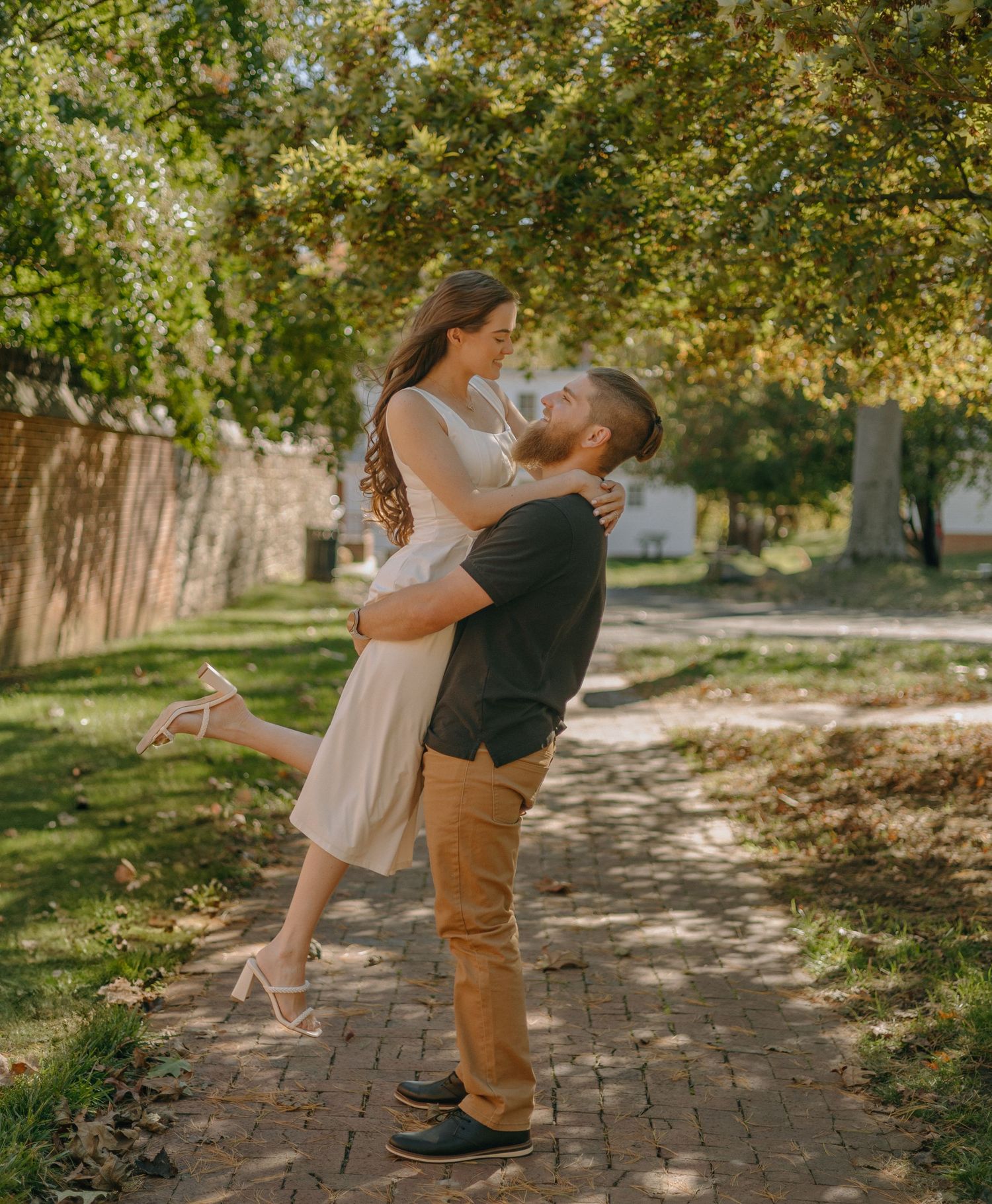 A couple embraces and dances together on a brick pathway surrounded by lush green trees in a romantic outdoor setting.