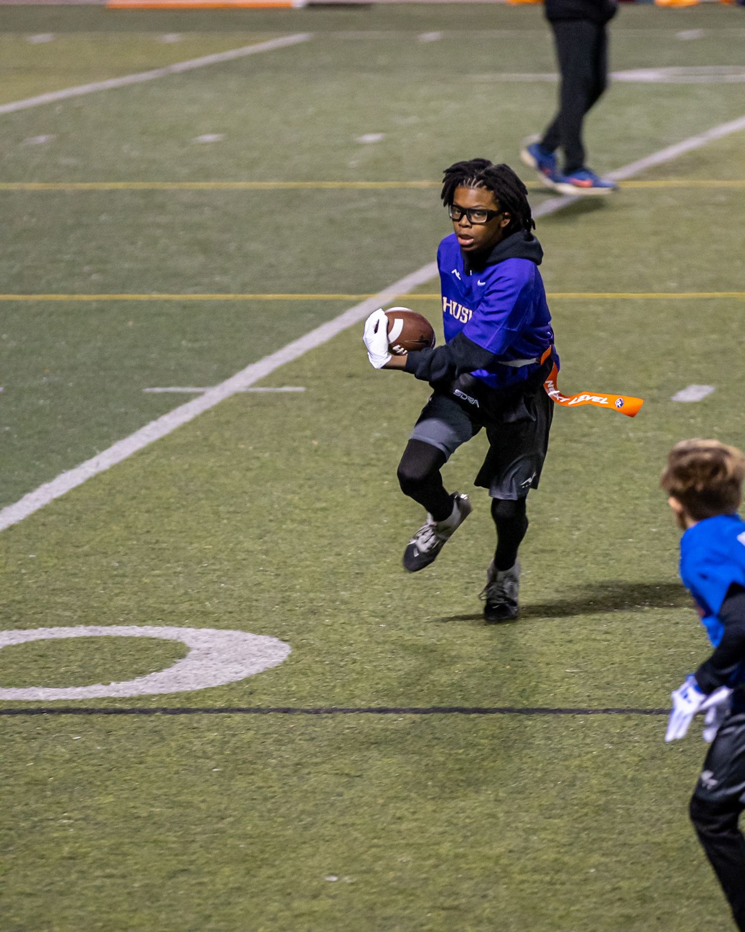 Player in blue uniform running with football on sports field at night.