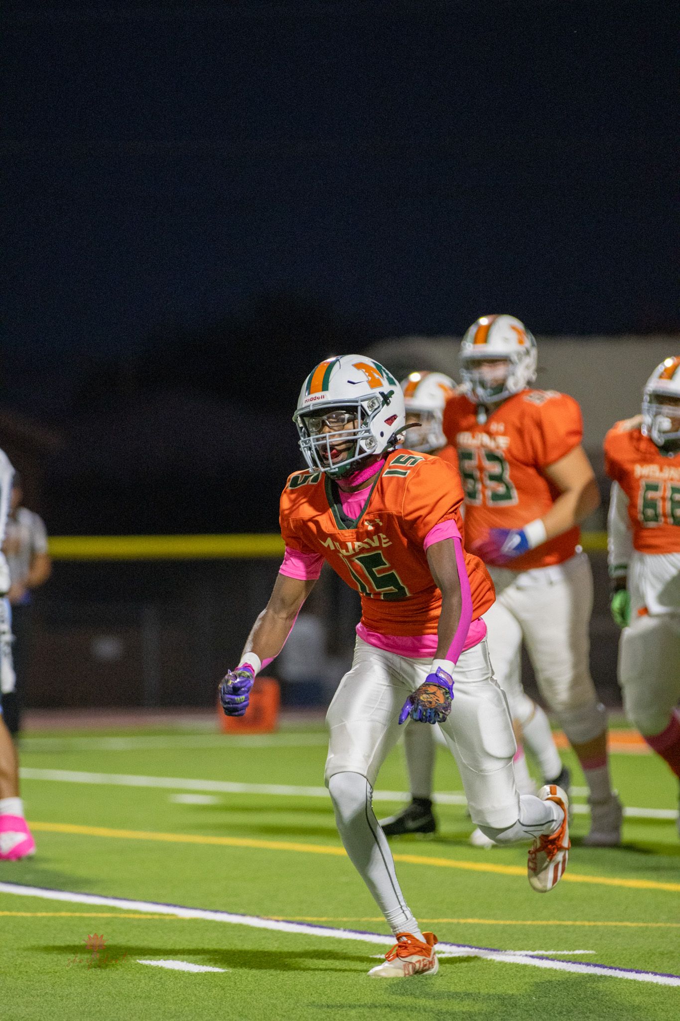 Football player in orange jersey and white pants running on field during night game.