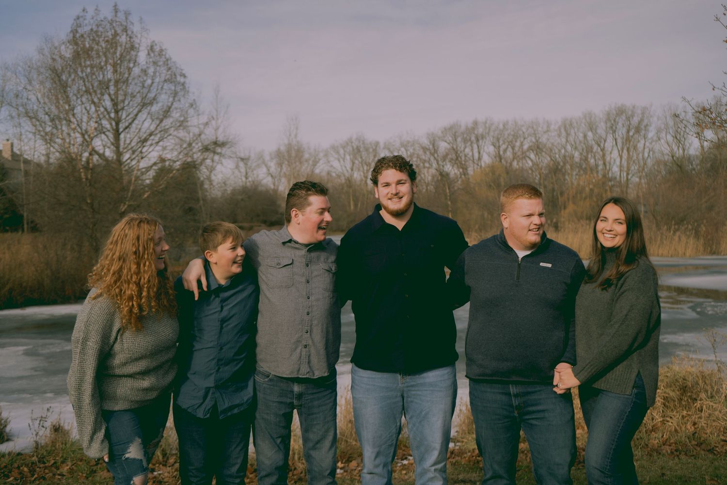 Five adults smiling and posing together outdoors near a frozen lake surrounded by bare winter trees.