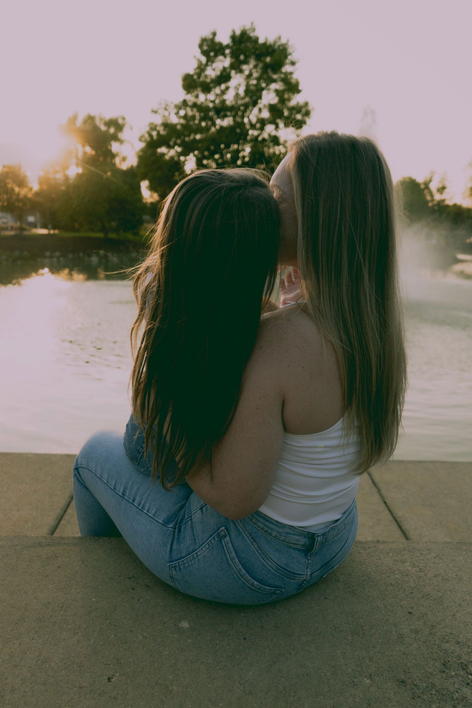 Two friends in matching blue jeans sit side by side watching the sunset at a waterfront location.