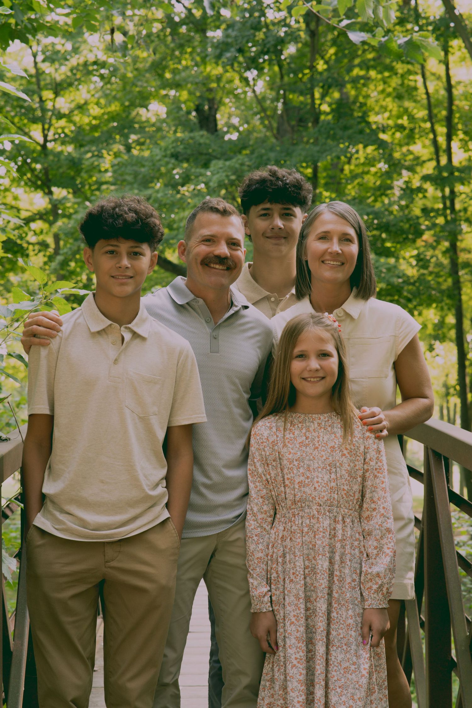 A group of people wearing light colored clothing stand together on a wooden deck surrounded by green trees.