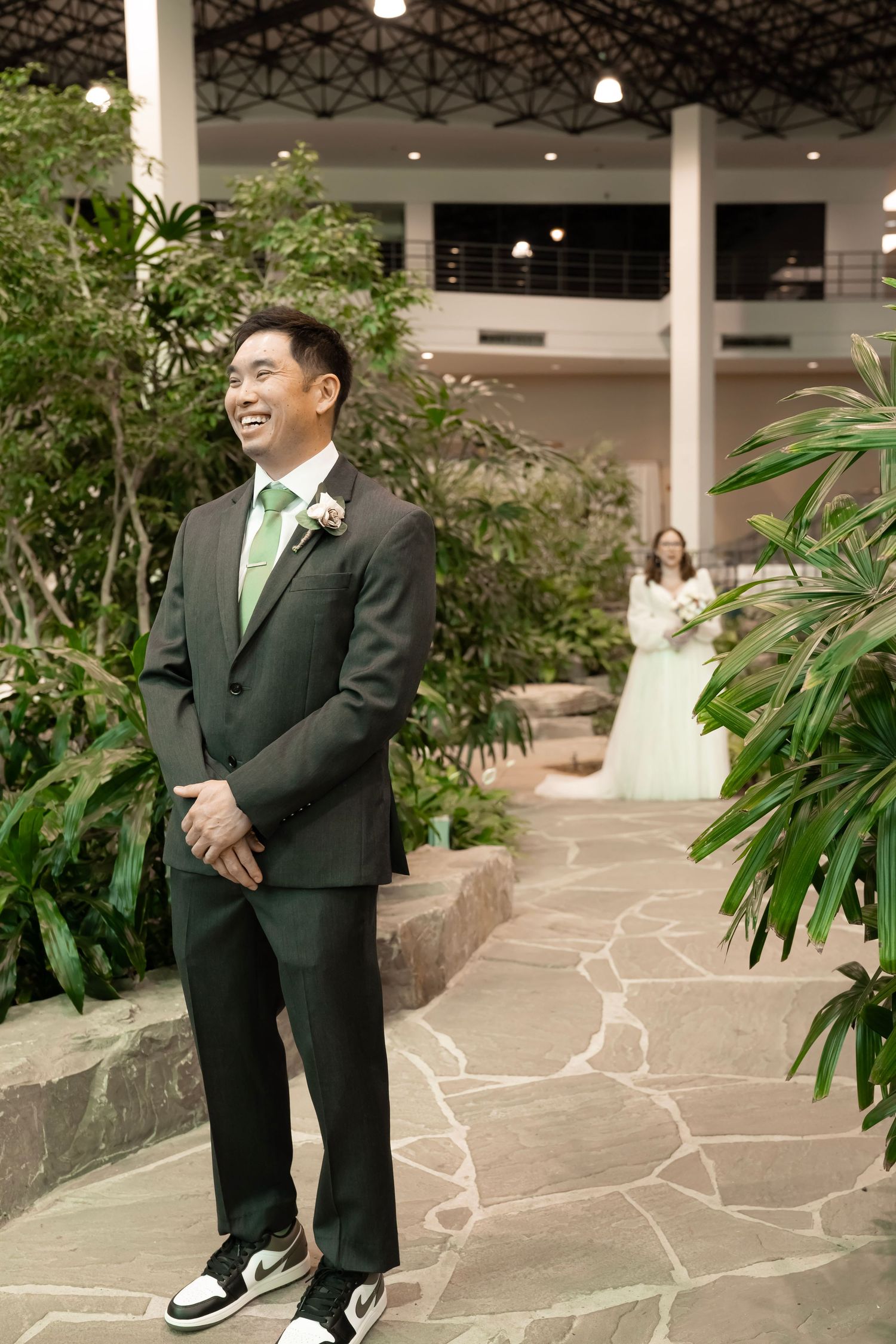 Groom in dark suit with green tie smiles as bride in white gown approaches during indoor wedding ceremony.