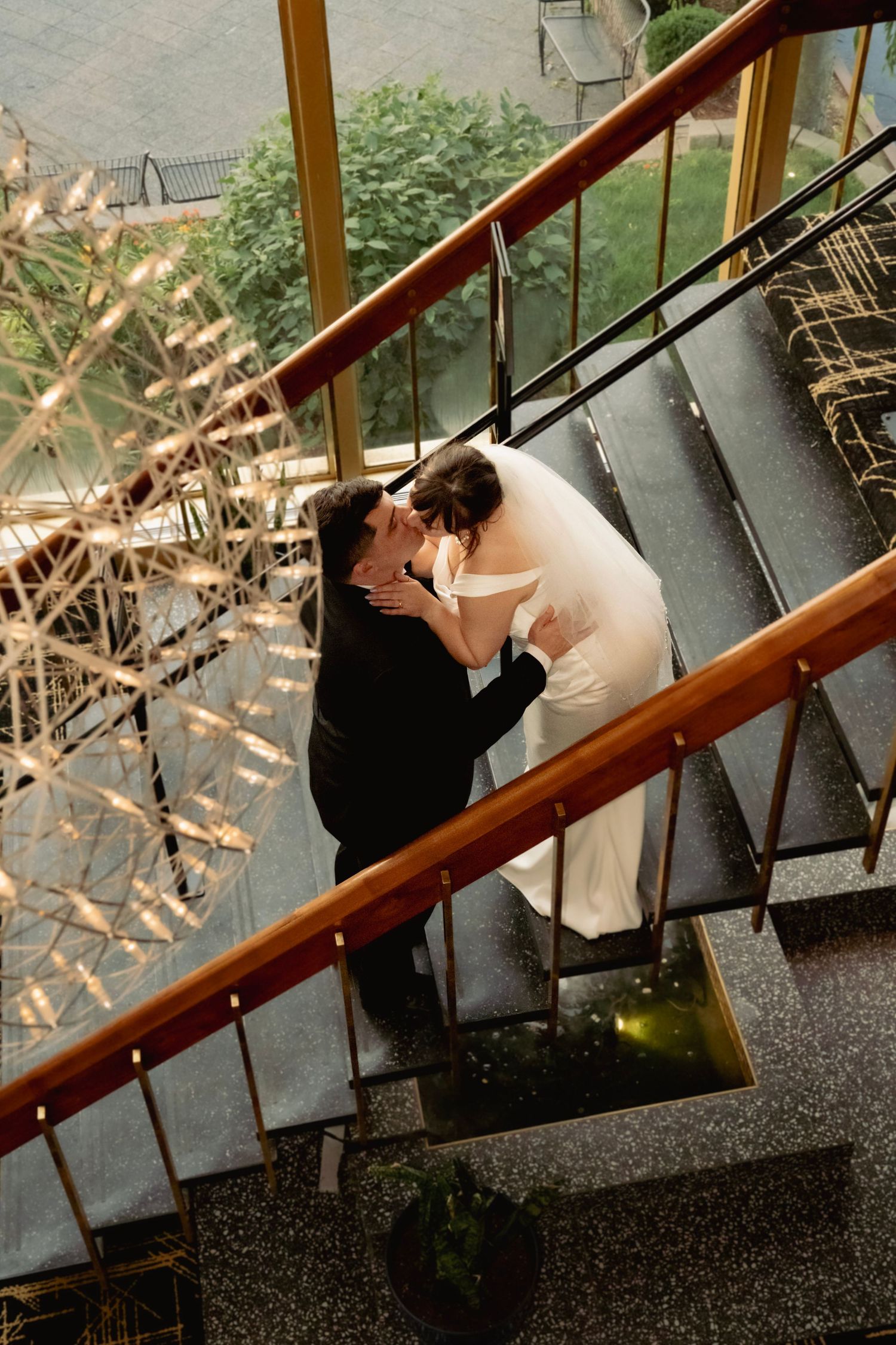 Aerial view of bride and groom sharing a kiss on elegant staircase with wood railings and chandelier at wedding venue.