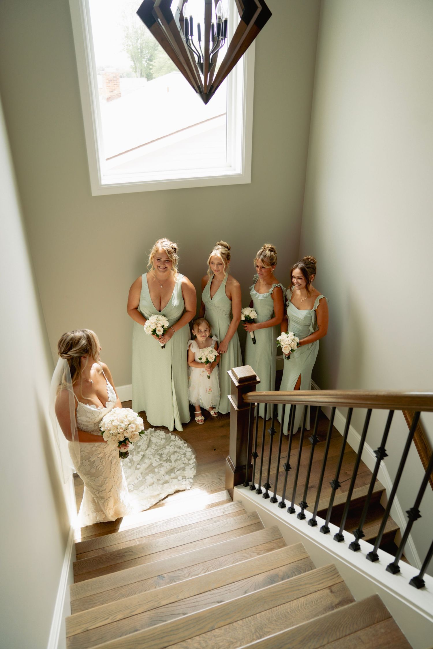 Bride in white dress and bridesmaids in mint green dresses pose joyfully on a wooden staircase with white walls.