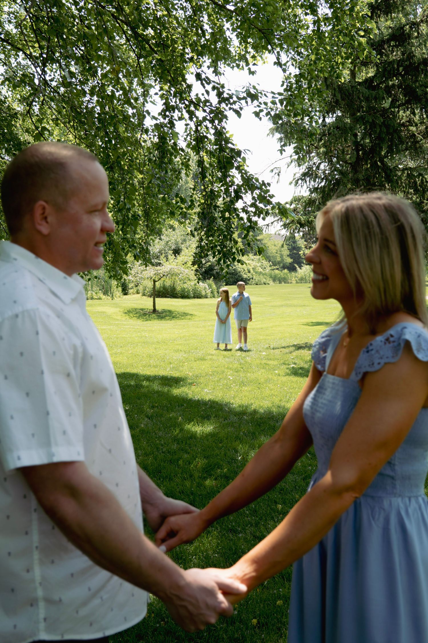 A couple holds hands in a romantic moment under green trees in a peaceful park setting.