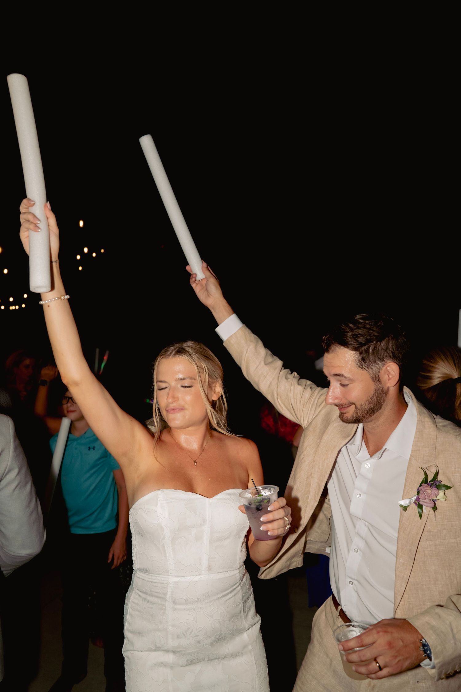 Bride and groom celebrating at outdoor wedding reception, raising glow sticks while holding cocktails at night.