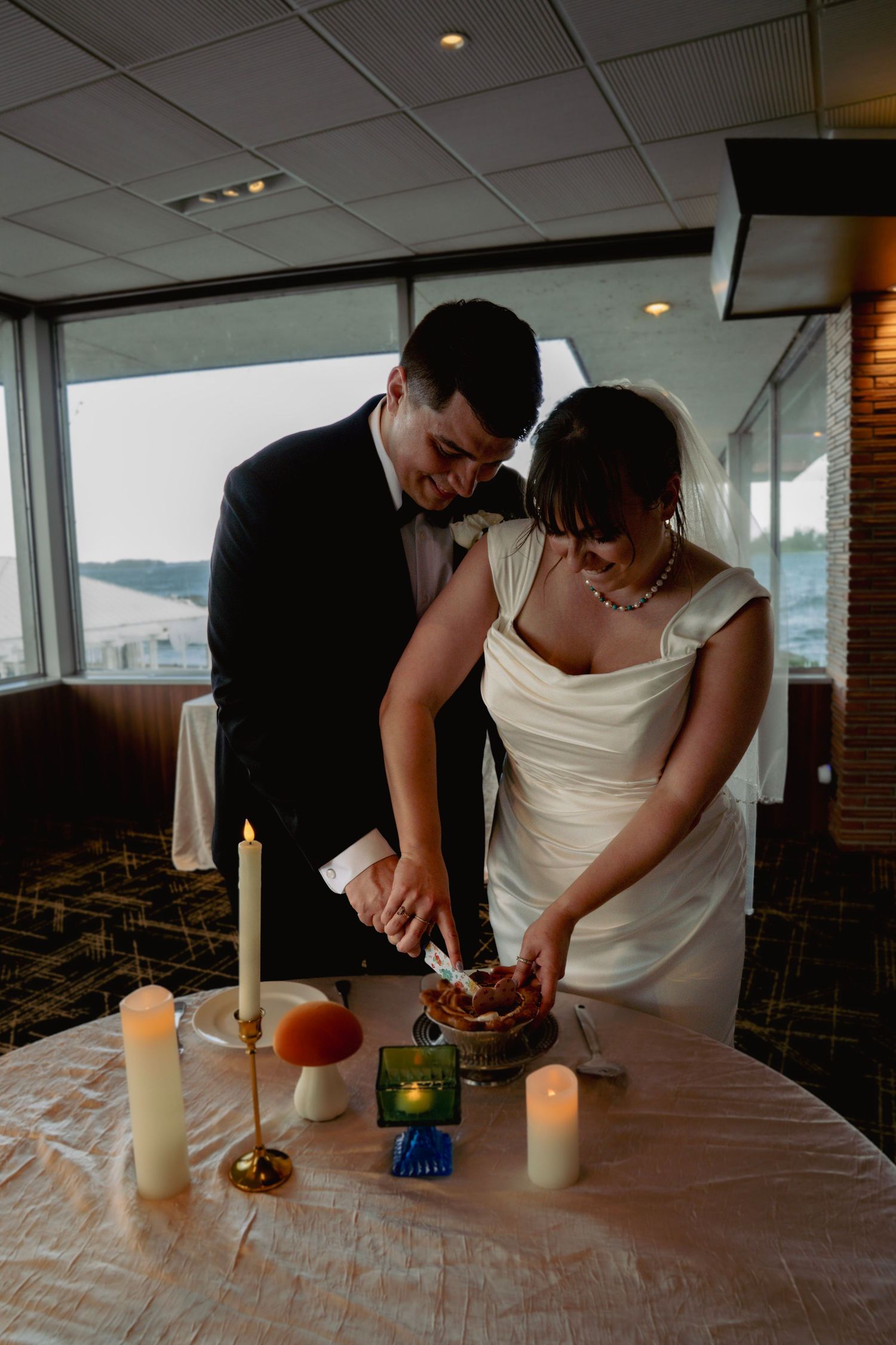 Bride and groom cutting wedding cake together at candlelit reception table with scenic waterfront view.