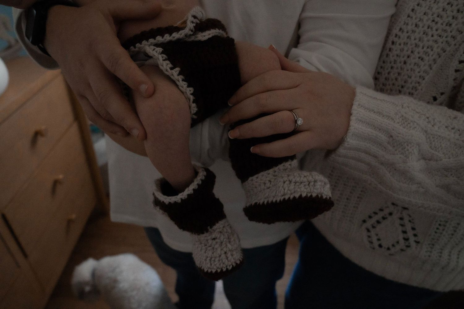 Couple holding tiny handmade crochet baby booties in black and cream, announcing pregnancy with wedding ring visible.