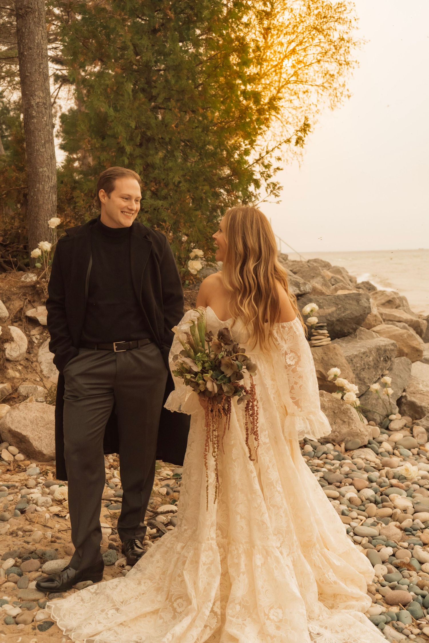 A romantic couple sits together on coastal rocks at sunset wearing black and cream-colored formal attire.